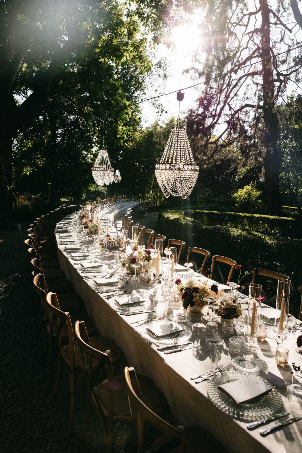 Outdoor wedding reception long table with crystal chandeliers hanging from trees at sunset.
