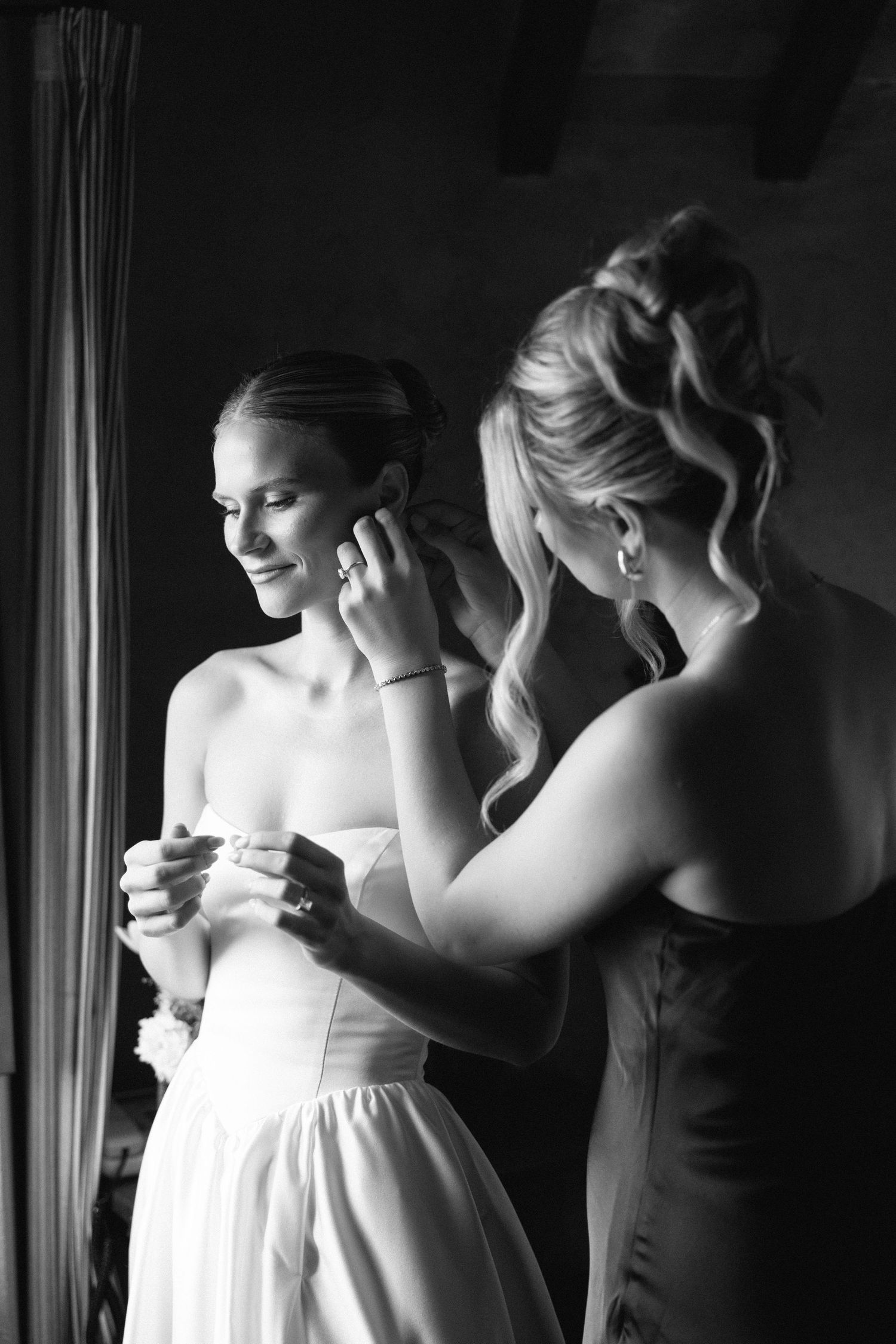 Black and white photo of a bridesmaid helping a smiling bride put on earrings while adjusting her strapless wedding dress.