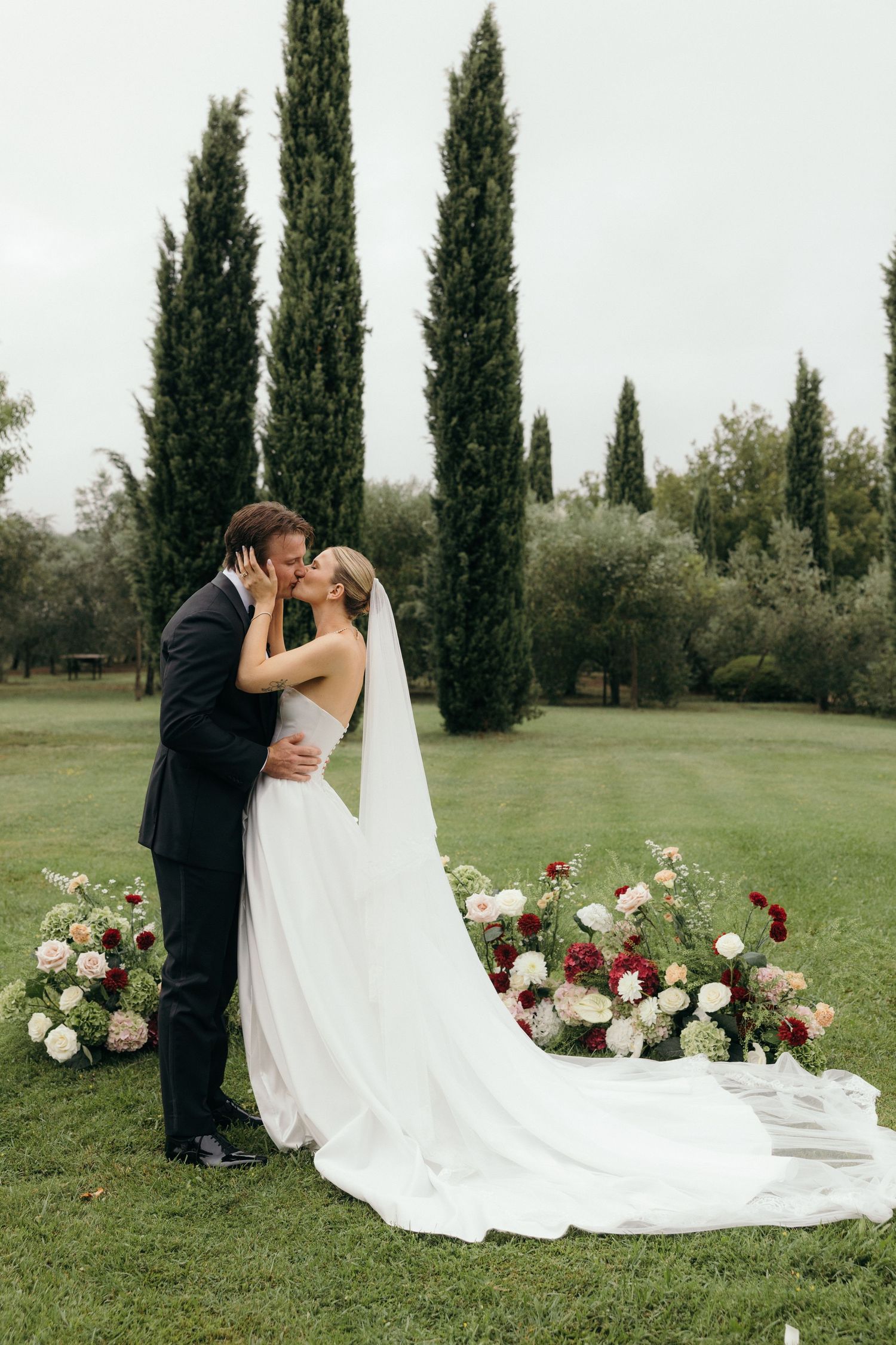 Bride and groom share a kiss outdoors at garden wedding ceremony with cypress trees and floral arrangements.