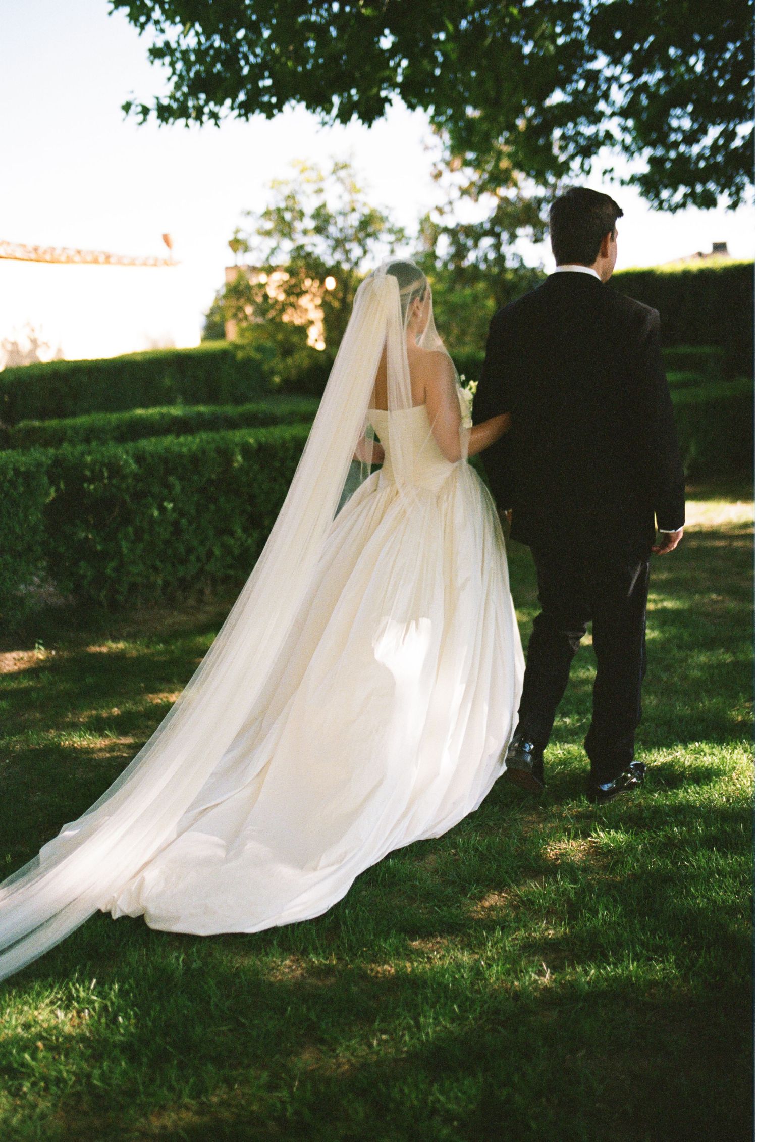 A bride in a flowing white gown with cathedral veil walks hand-in-hand with her groom through a lush garden.