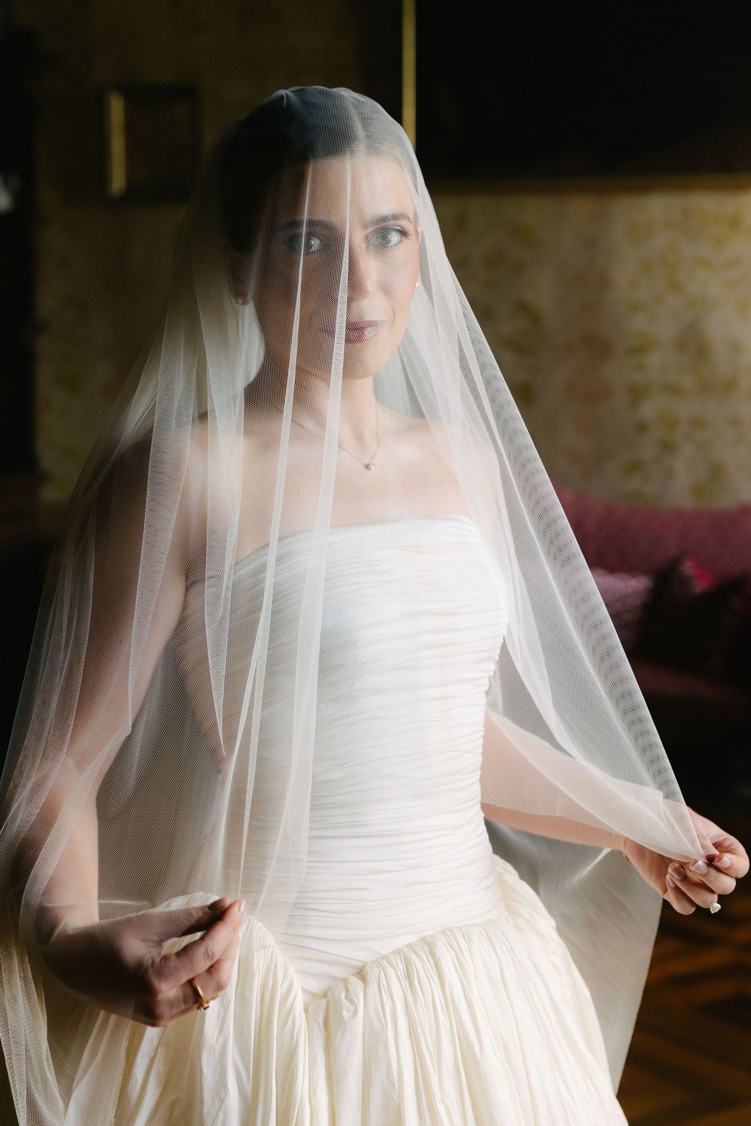 Beautiful bride in strapless white gown holding sheer veil over face in a softly lit elegant room.