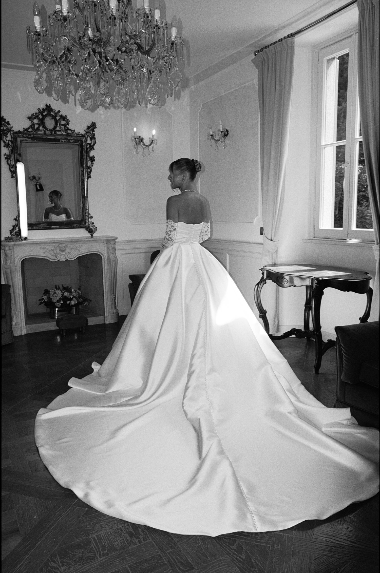 Bride in elegant off-shoulder ball gown with long train stands in ornate room beneath crystal chandelier, black and white photo.
