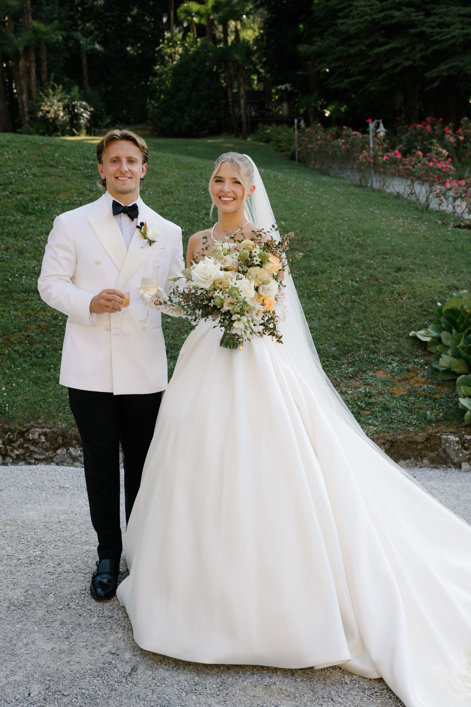 Bride in white ball gown holding floral bouquet stands with groom in white tuxedo jacket outdoors on wedding day.