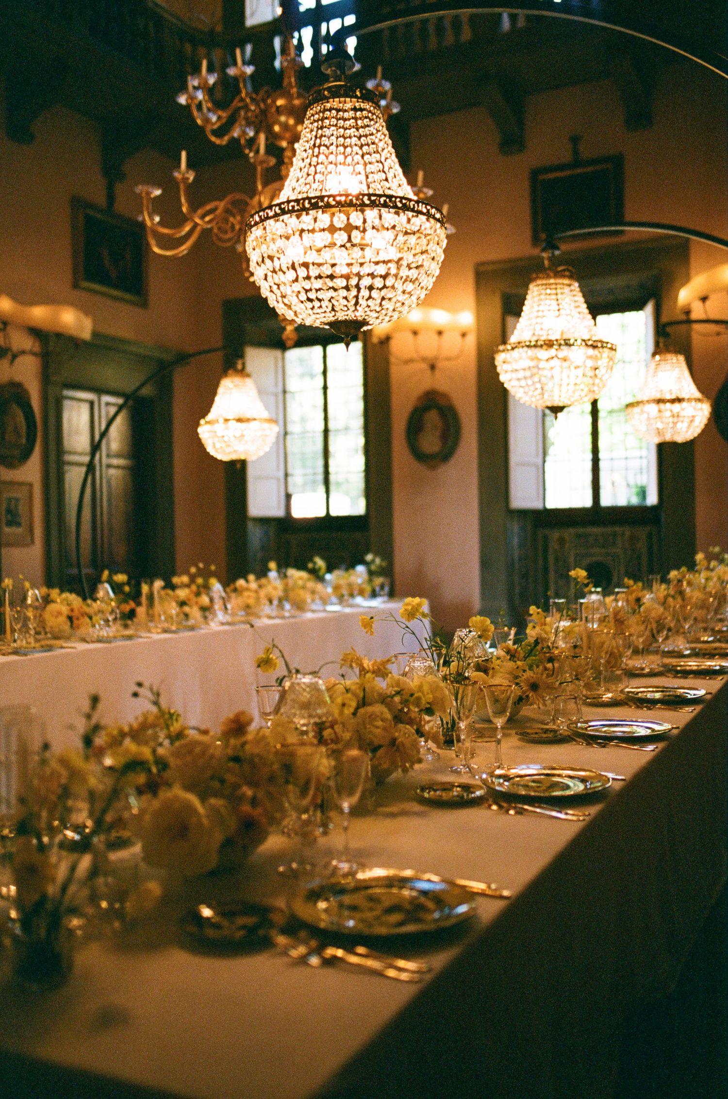Luxurious wedding reception table set with crystal chandeliers, white florals, and gold place settings in ornate hall.