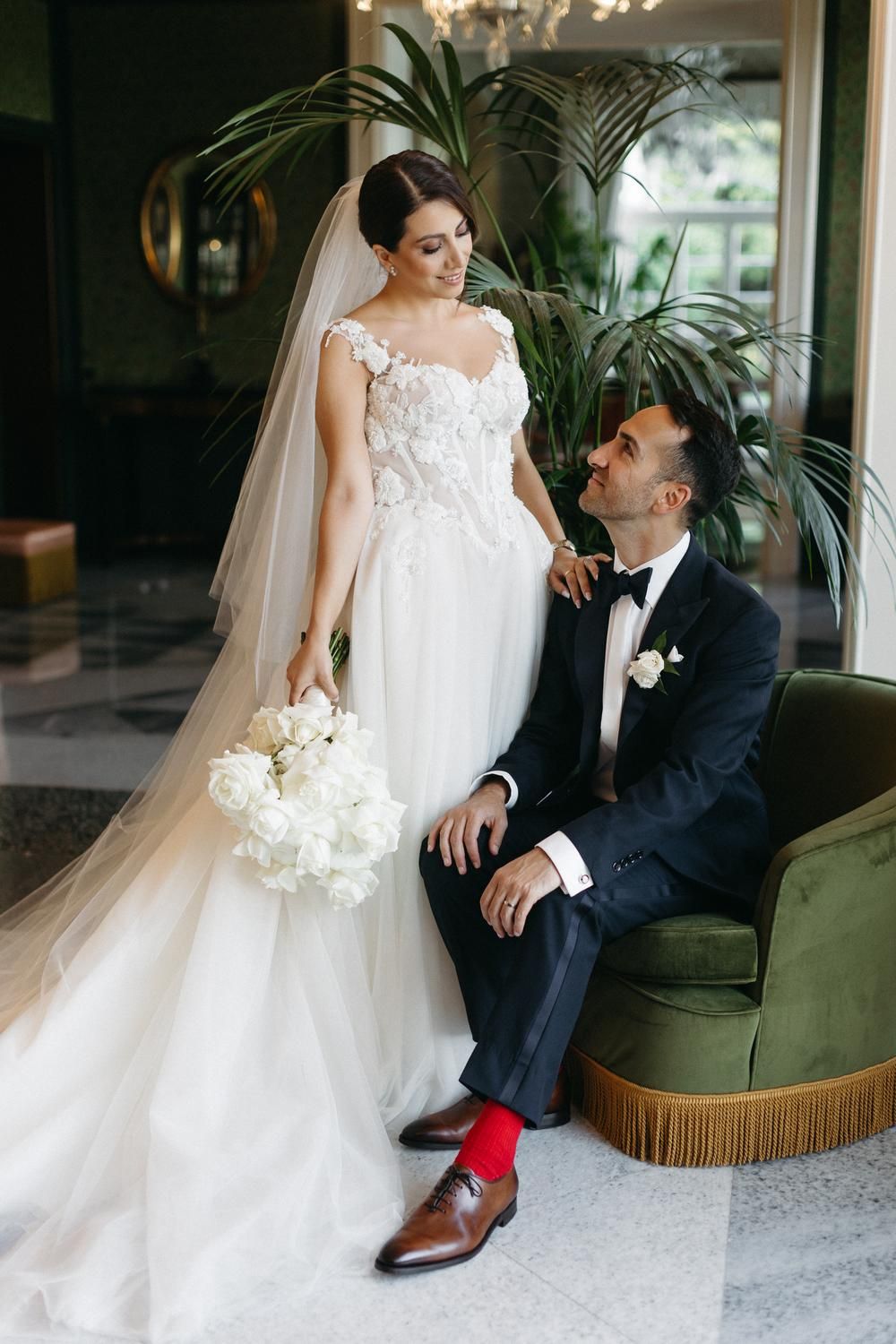 Bride in floral gown holding white bouquet stands beside groom in navy tuxedo with red socks on green velvet chair.