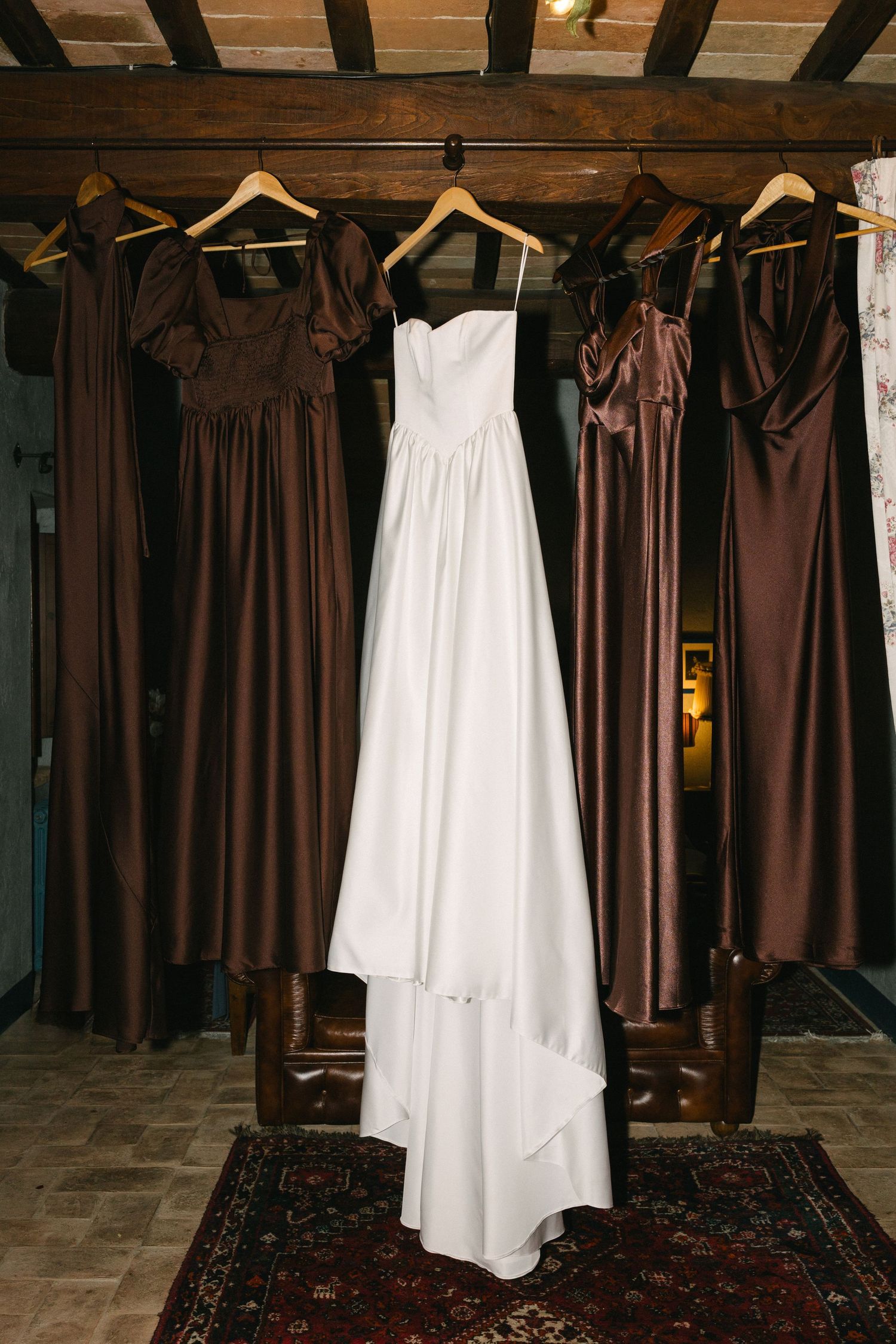 White wedding gown and brown satin bridesmaid dresses hanging on wooden hangers in a rustic venue.