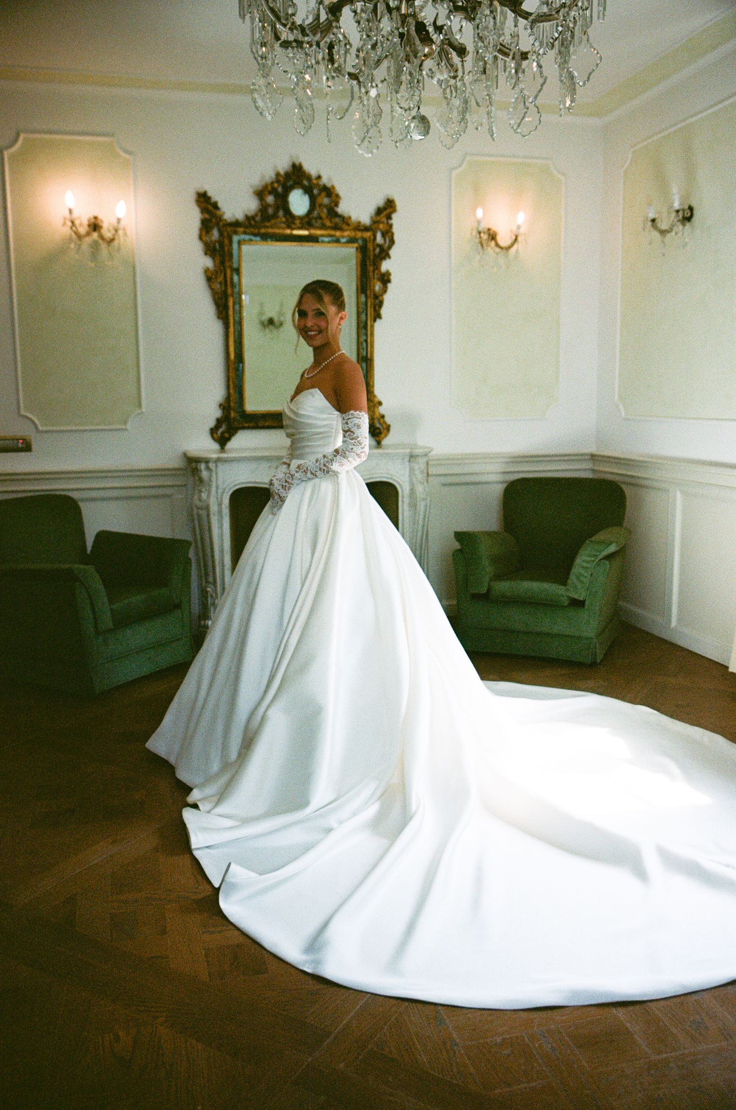 Bride in elegant white ball gown with long train poses in ornate room with crystal chandelier and gold mirror.
