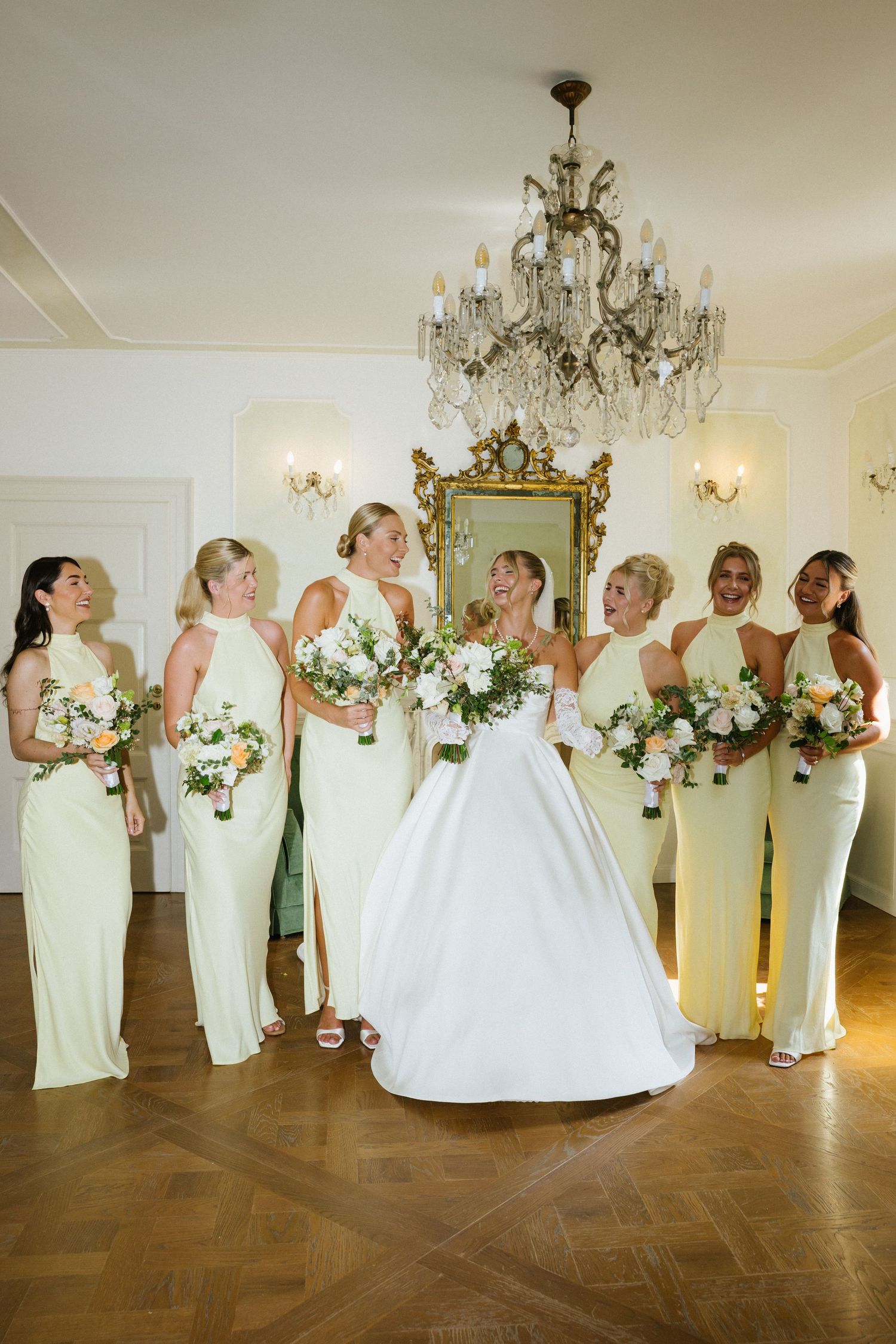 Bride in white ball gown poses with six bridesmaids in pale green dresses holding bouquets under a crystal chandelier.