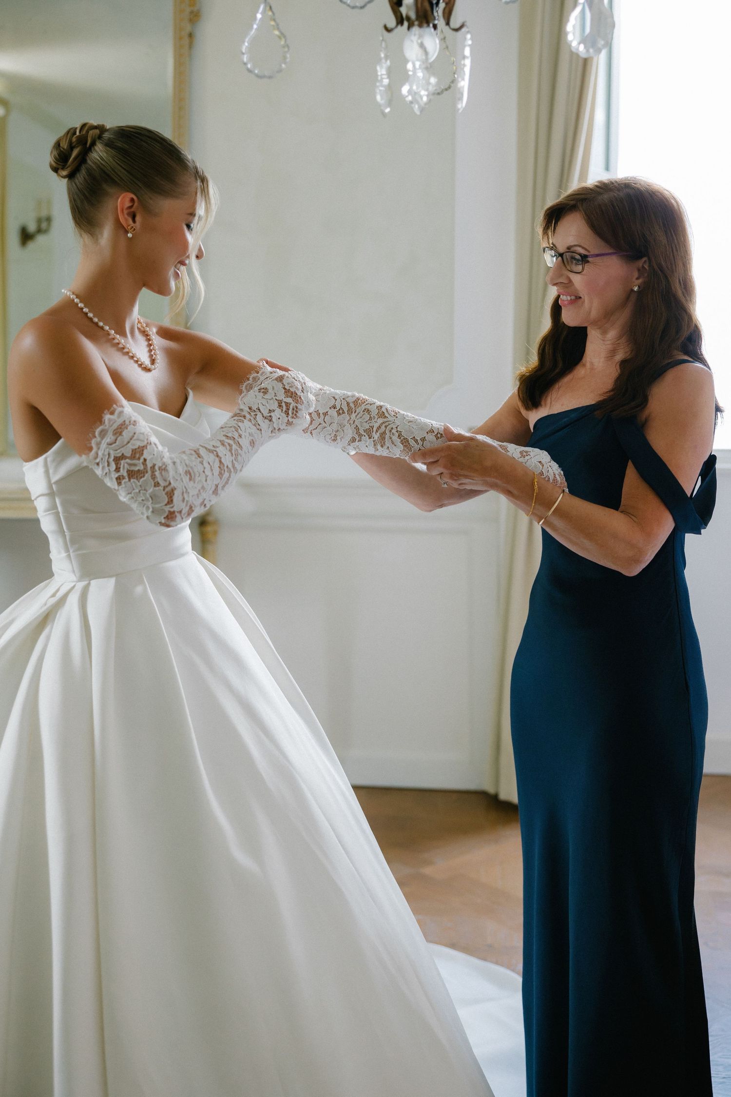Bride in white lace gloves and ball gown holds hands with woman in navy dress during wedding preparation.