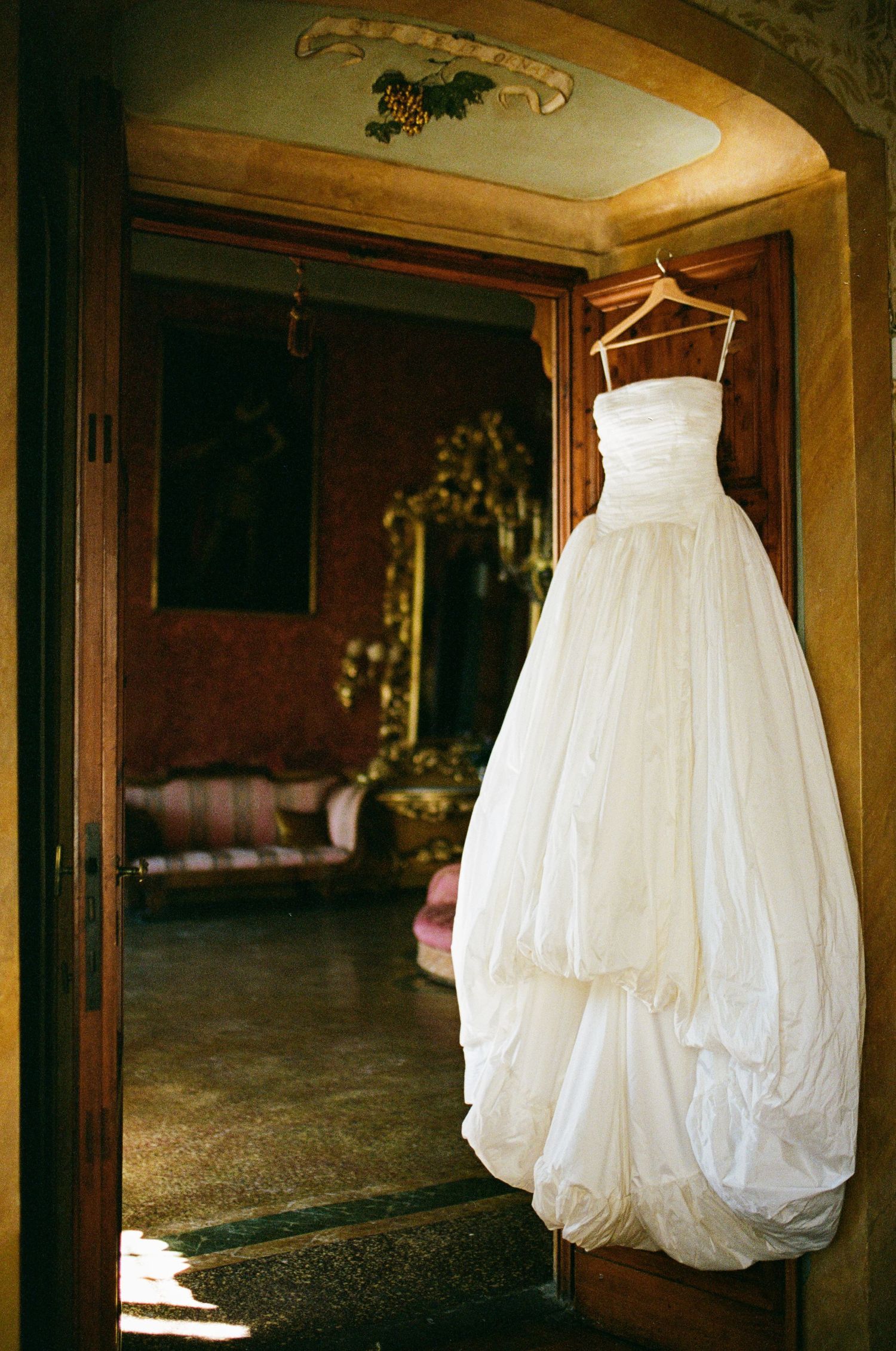 Strapless white ball gown wedding dress hanging on wooden door in ornate vintage Italian villa room.