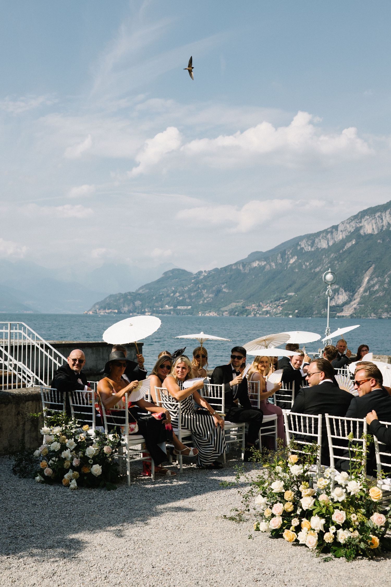 Outdoor wedding ceremony by Lake Como with guests seated on chiavari chairs, white parasols, floral arrangements, and mountain views.