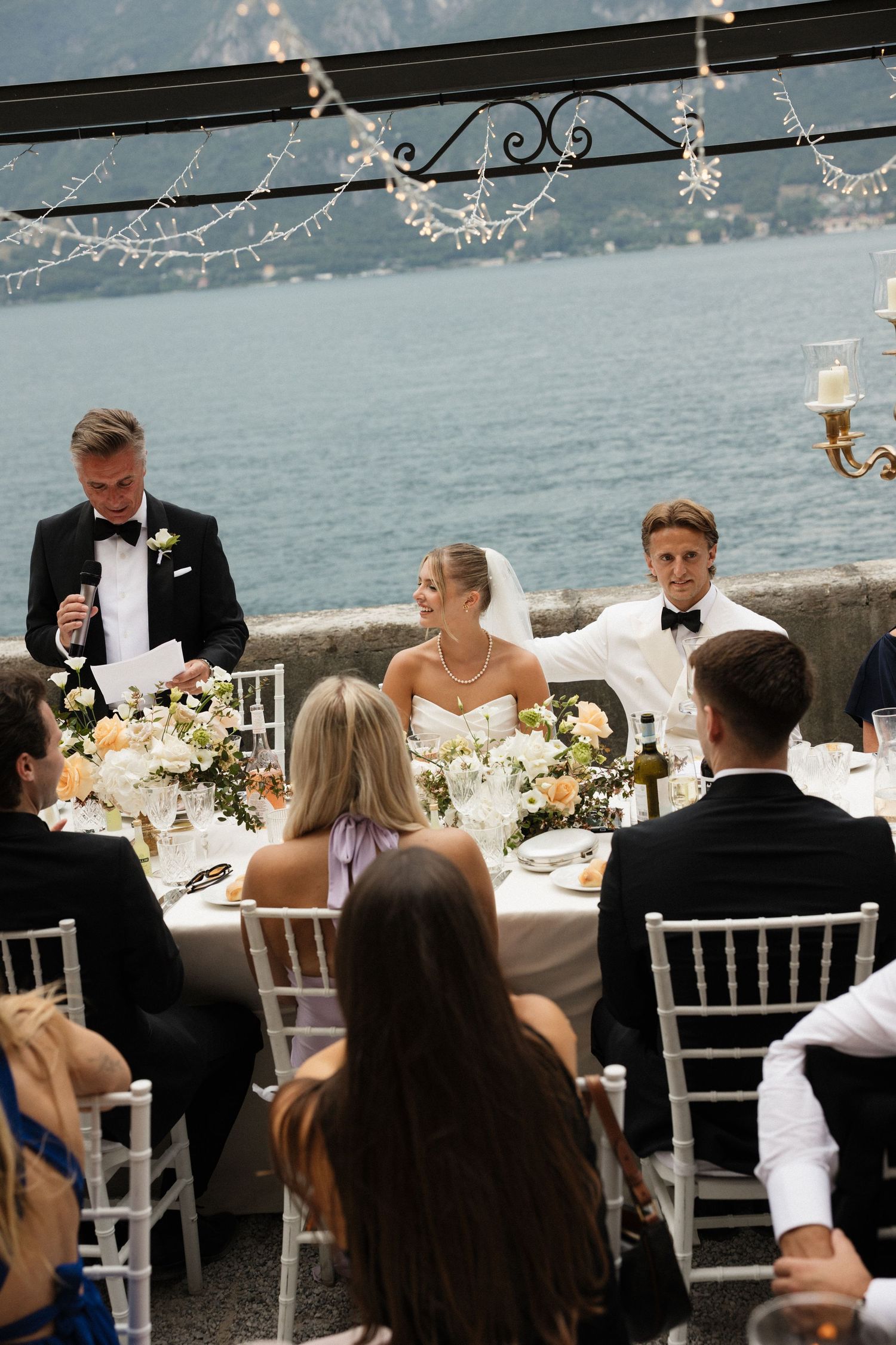 Elegant wedding reception on Lake Como terrace with bride and groom at decorated table, guests seated, fairy lights overhead.