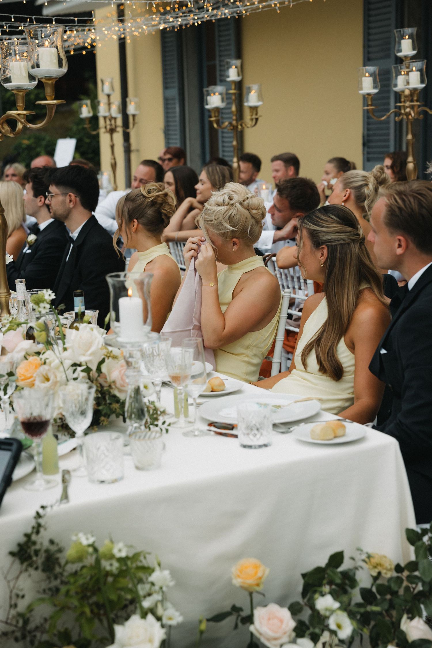 Elegant outdoor wedding reception with guests in formal attire seated at flower-adorned tables with white linens.