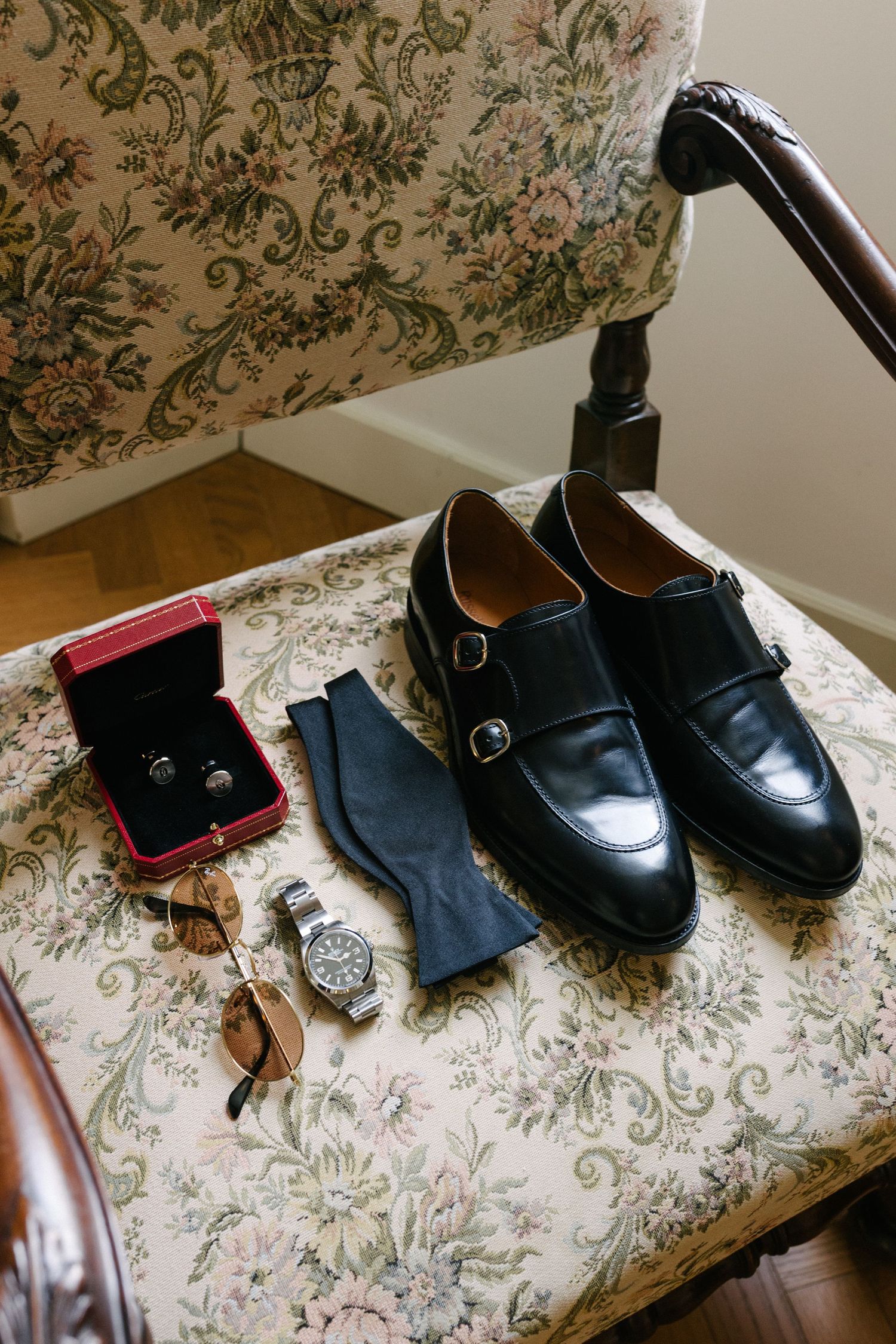 Black monk strap dress shoes with cufflinks, bow tie, watch, and sunglasses on a floral upholstered chair.