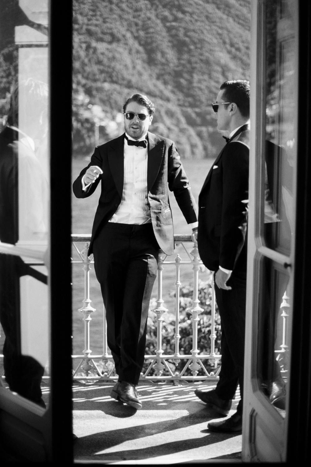 Black and white photo of two groomsmen in tuxedos with sunglasses laughing on a lakeside balcony.