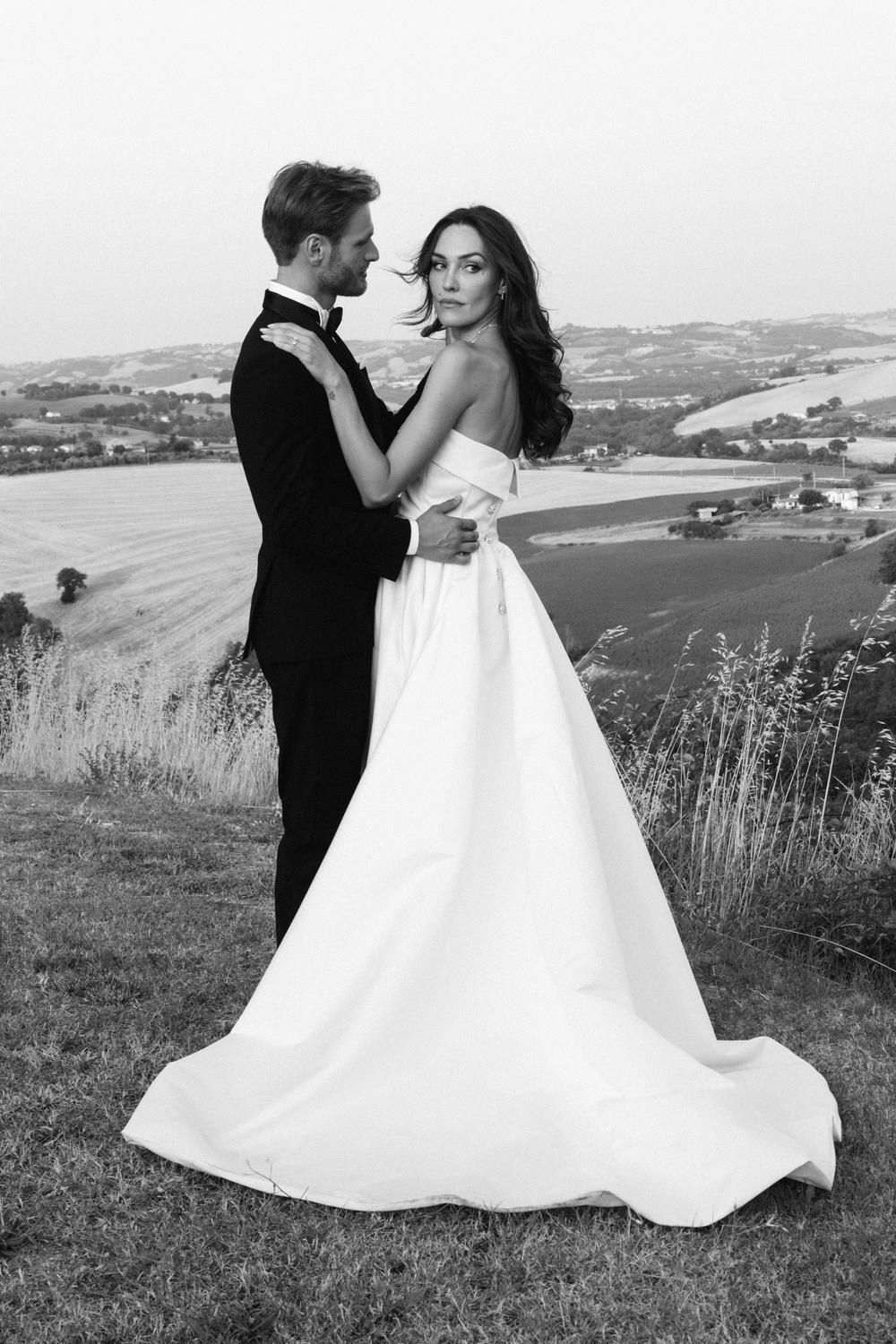 Black and white photo of a bride and groom embracing on a hilltop with scenic countryside views behind them.