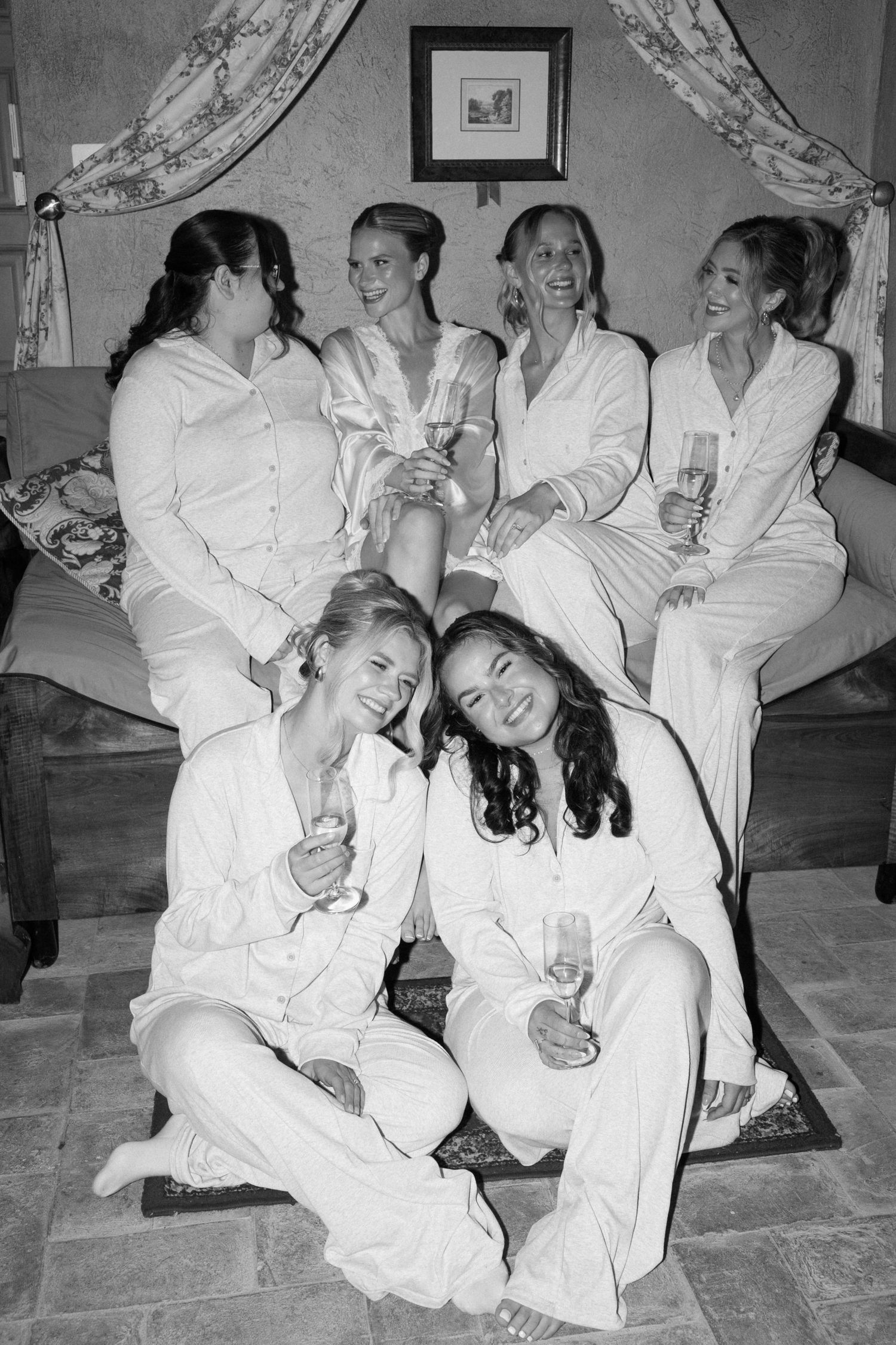 Black and white photo of six bridesmaids in white pajamas toasting with champagne glasses before a wedding ceremony.