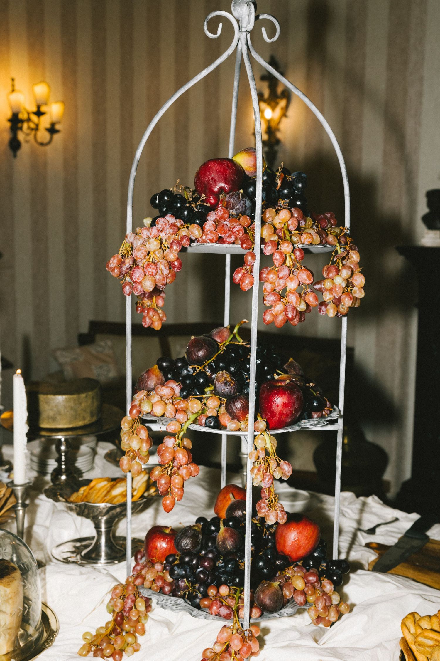 Elegant white metal tiered stand displaying fresh red and black grapes with apples, styled as a decorative fruit display.