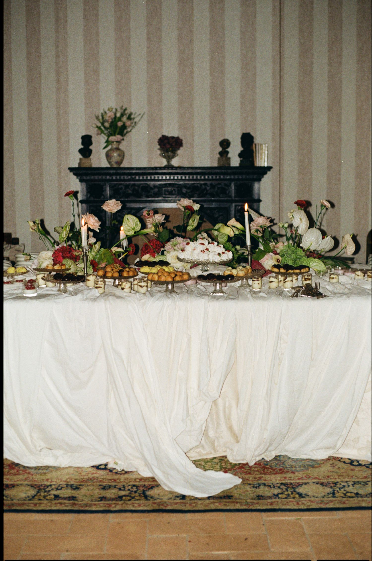Elegant banquet table with white draped cloth, floral arrangements, candles, and assorted foods near a dark fireplace.