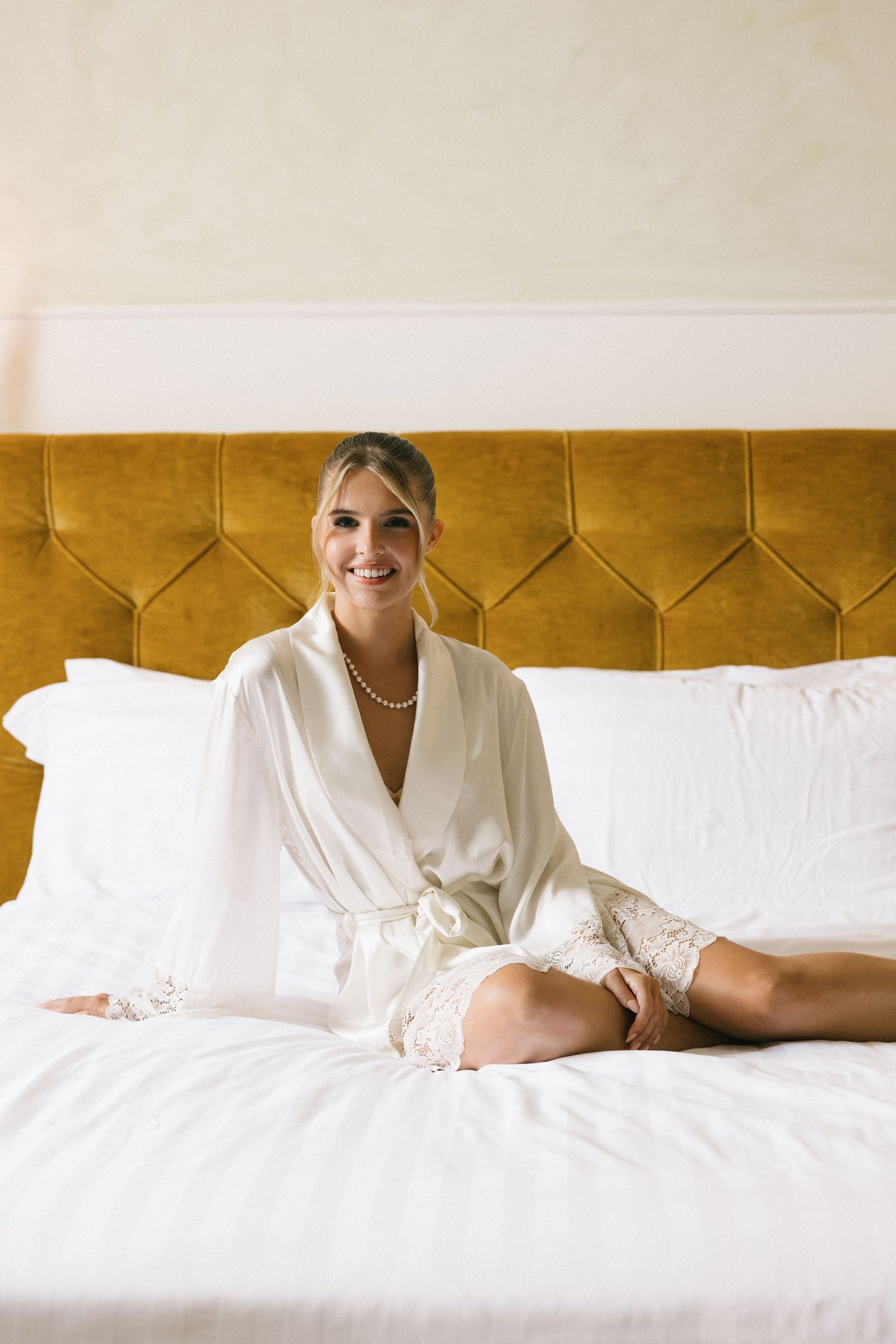 Smiling bride in white satin robe and pearl necklace sitting on a bed with white linens and gold headboard.