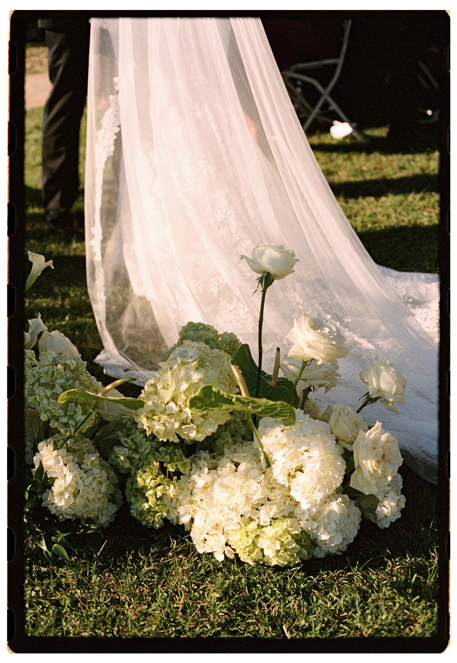 White hydrangeas and roses rest on grass beside a flowing bridal gown train at an outdoor wedding ceremony.