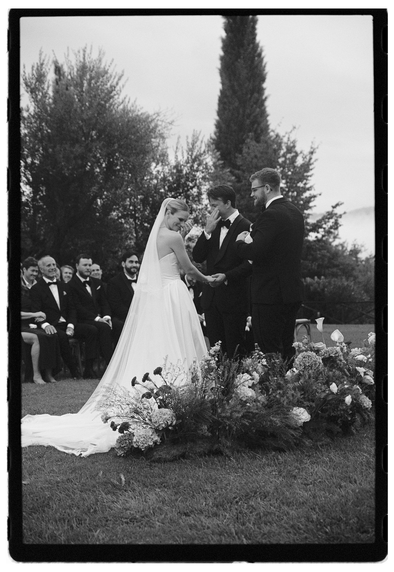 Black and white outdoor wedding ceremony with bride in veil, groom, officiant, and floral arrangement, guests seated in background.