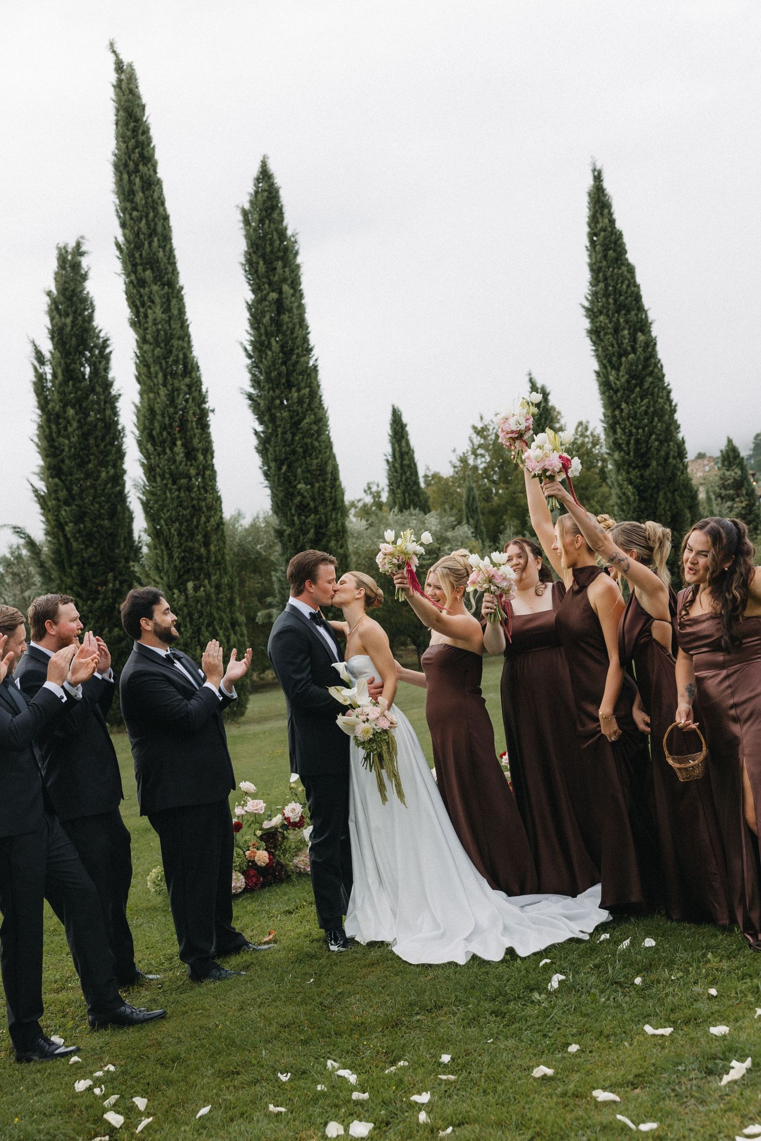 Bride and groom share a kiss while bridesmaids in brown gowns celebrate with bouquets at an outdoor wedding ceremony.