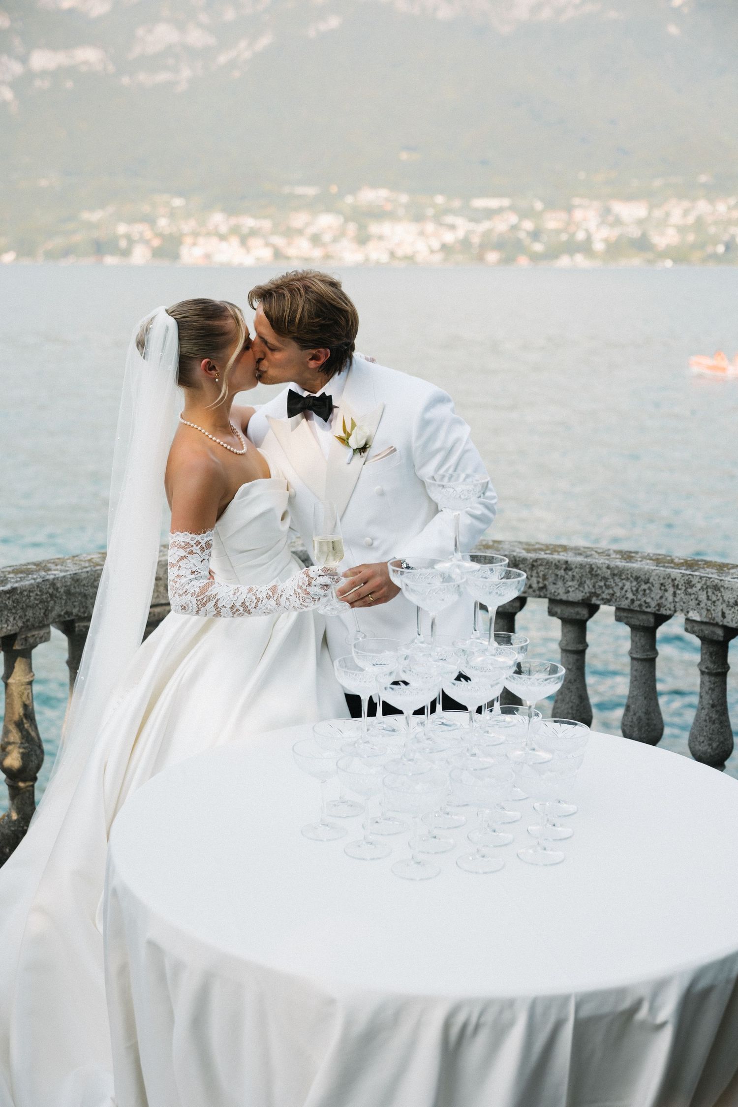 Bride and groom kissing beside champagne glasses tower on lakeside terrace with mountain views.