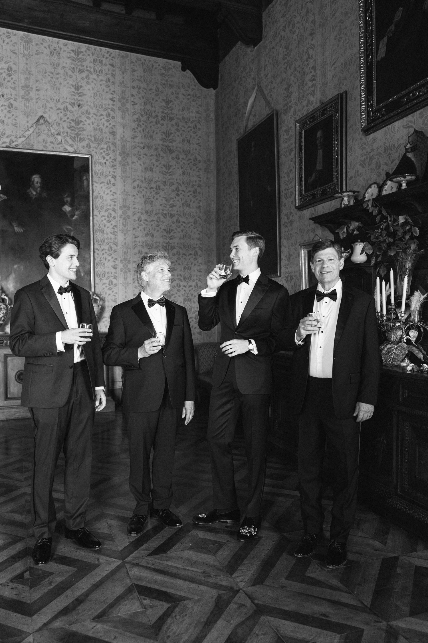 Four men in tuxedos toasting with drinks in an elegant room with ornate wallpaper and classic paintings.