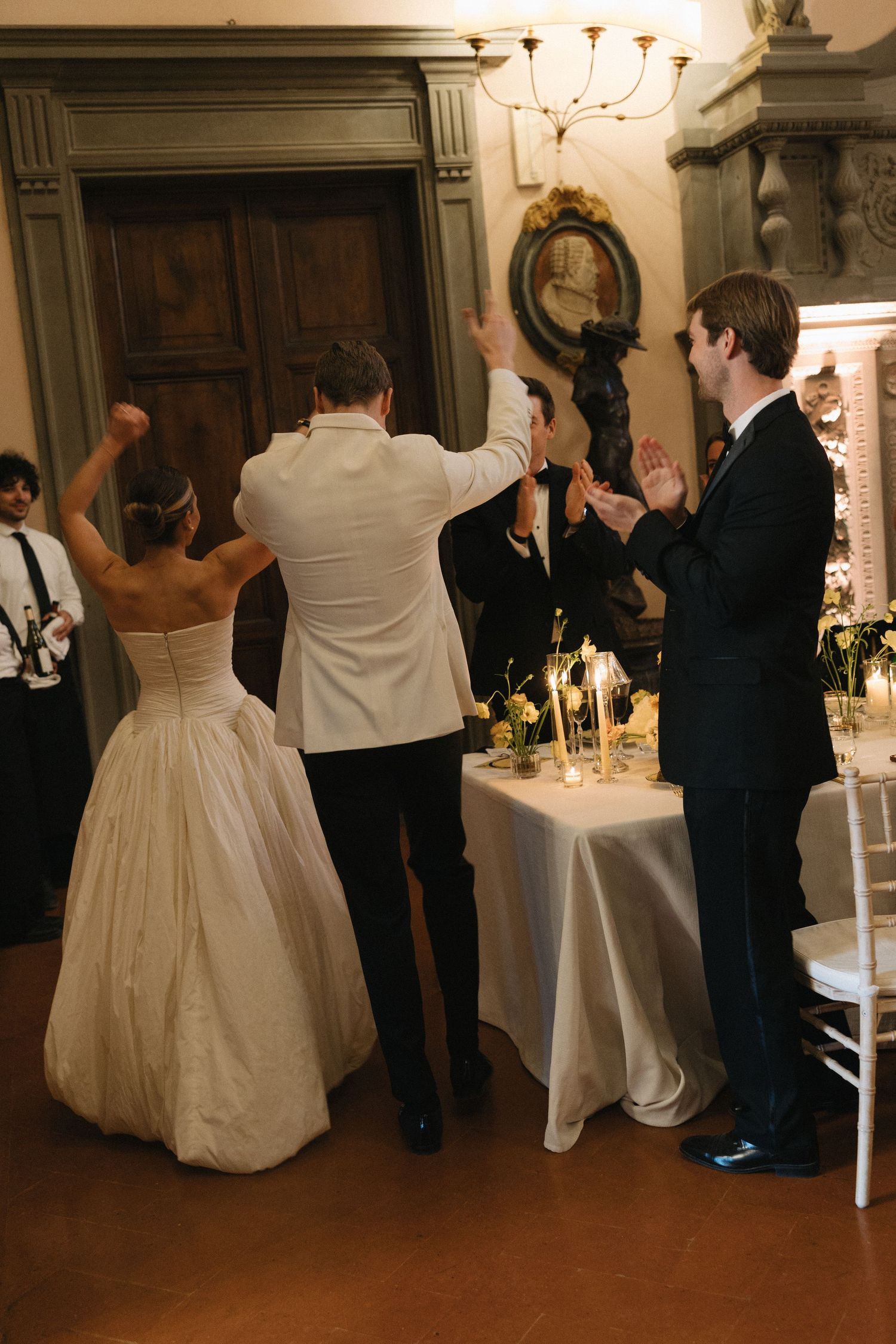 Bride and groom celebrate with raised arms at elegant wedding reception in ornate venue with candles.