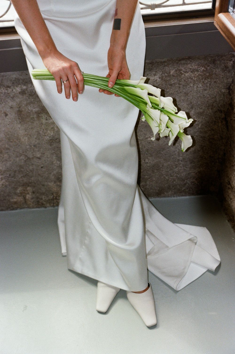 Bride in sleek white satin gown holding calla lily bouquet with white square-toe heels.