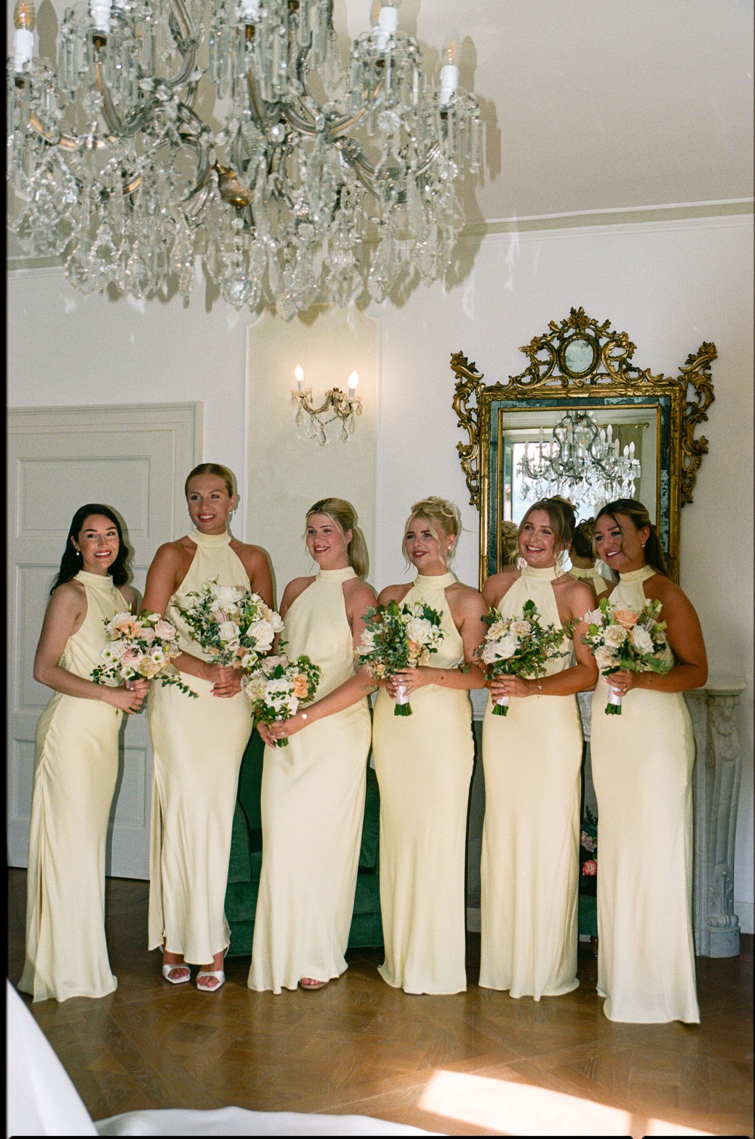 Six bridesmaids in matching yellow halter gowns holding floral bouquets beneath a crystal chandelier in an elegant venue.