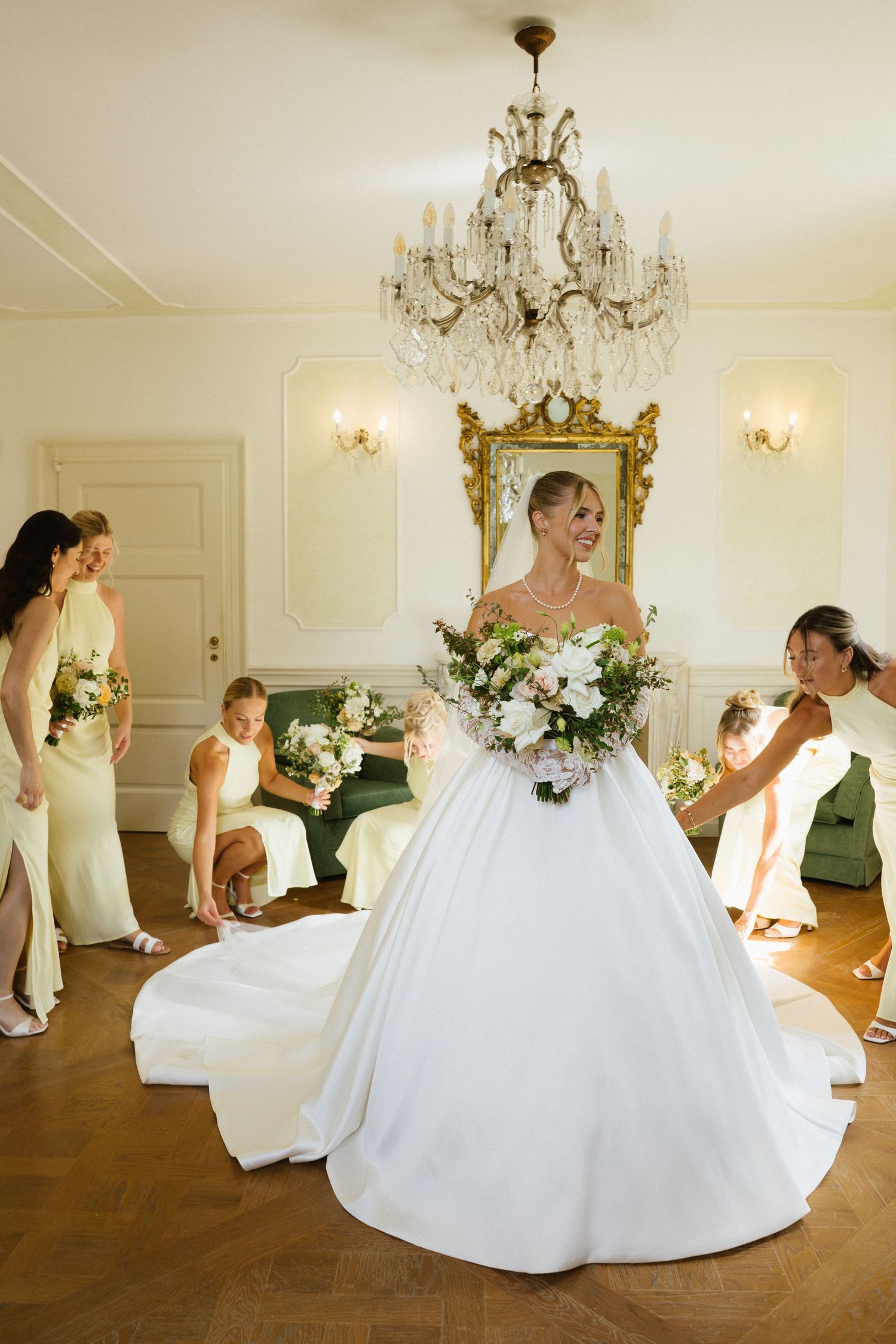 Bride in white ballgown holds floral bouquet surrounded by bridesmaids in sage green dresses in elegant chandelier room.