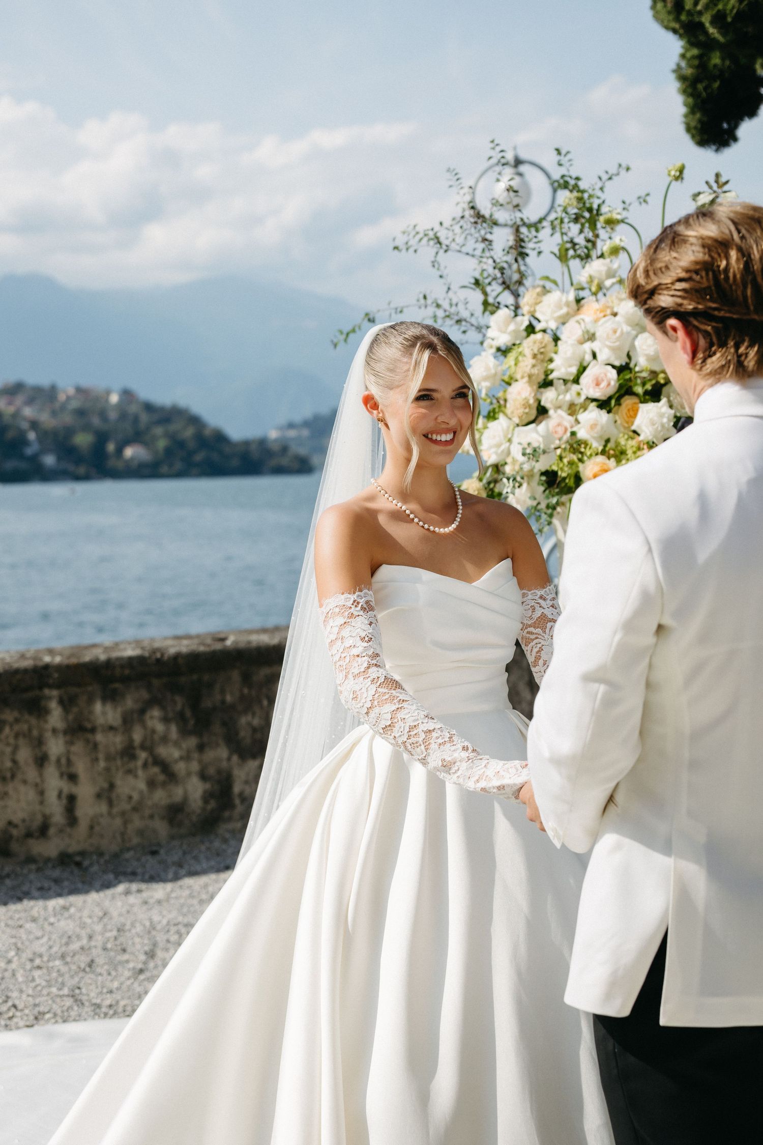 Bride in strapless white gown with lace gloves smiles at groom during outdoor lakeside wedding ceremony.
