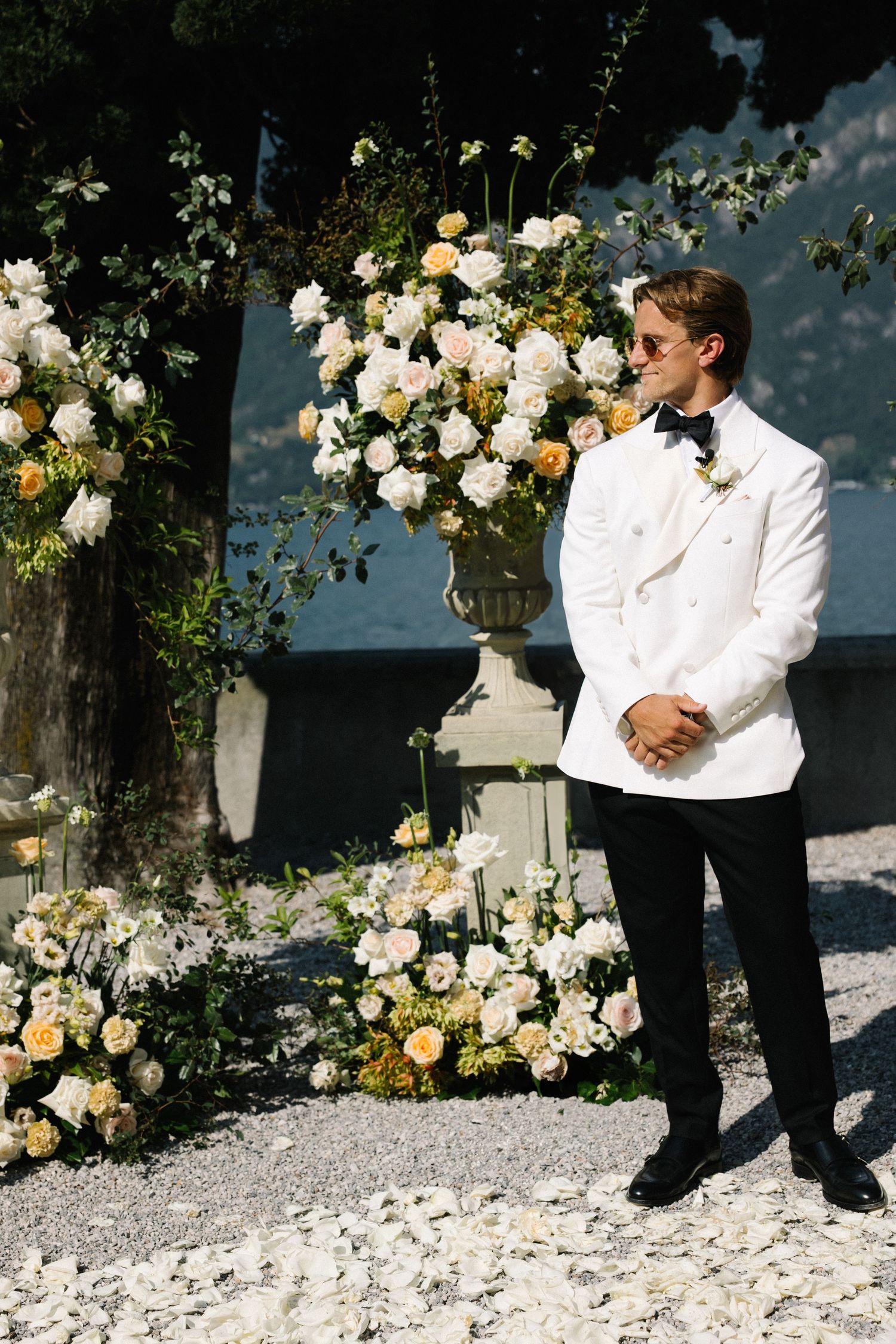 Groom in white tuxedo jacket stands before elegant floral arrangements of white and peach roses at outdoor wedding.