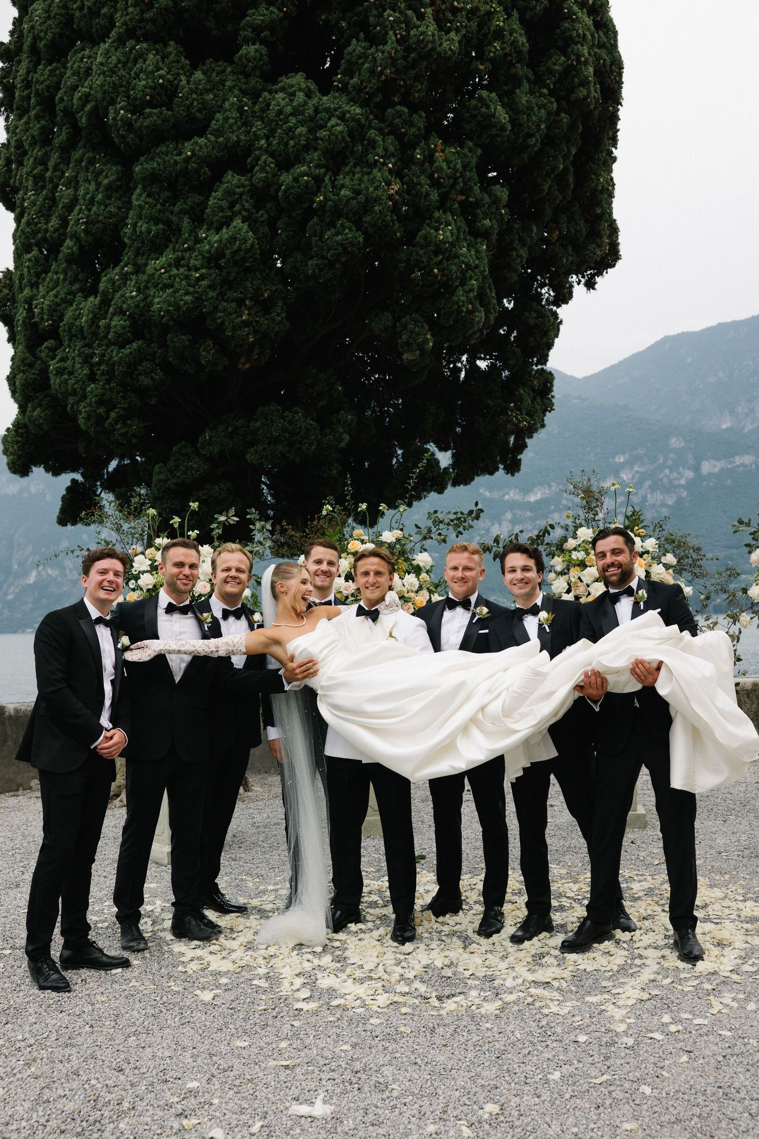 Groomsmen in black tuxedos lift smiling bride in white gown outdoors with mountain lake backdrop at elegant wedding.