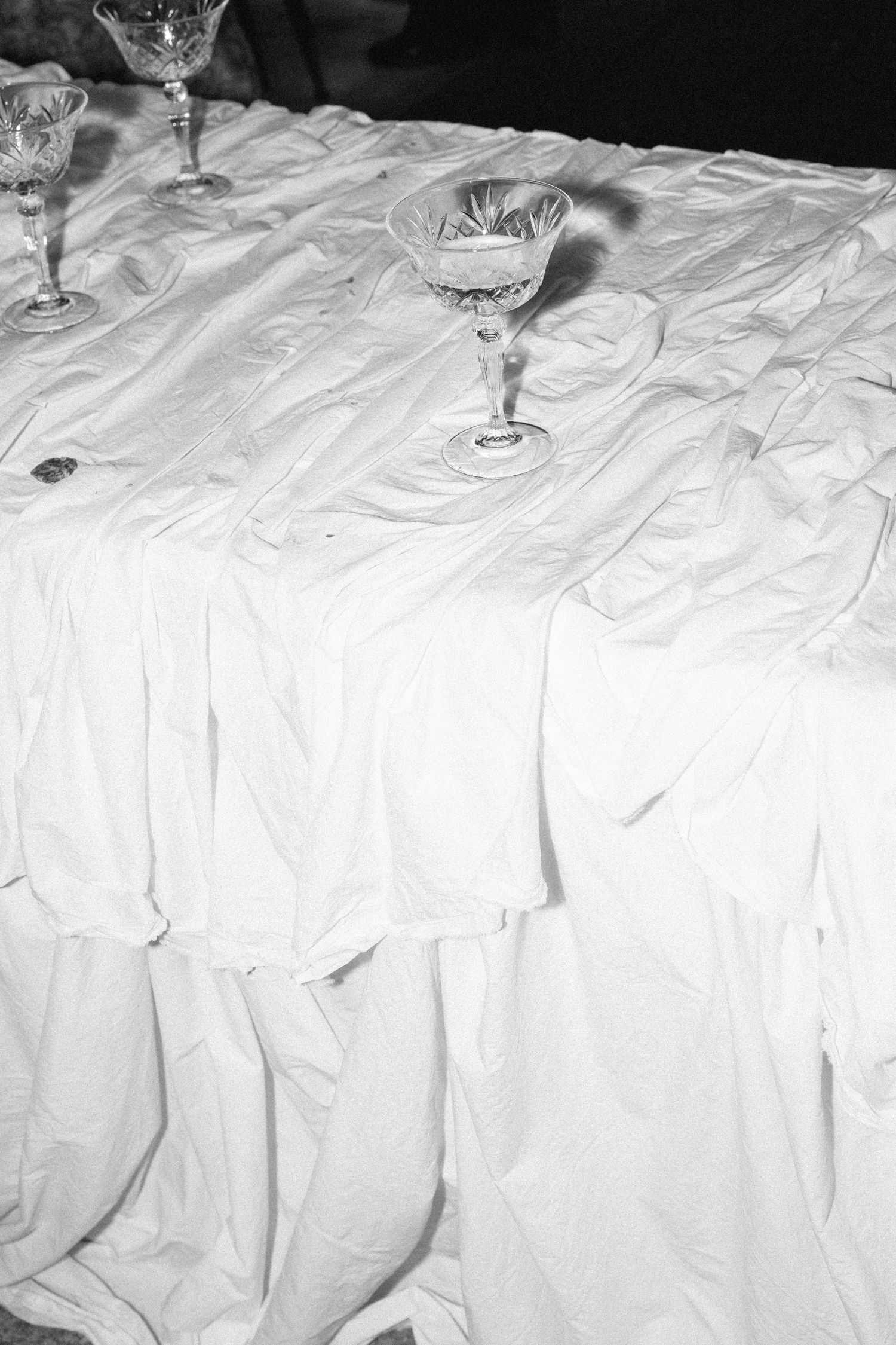 Black and white photo of crystal wine glasses on a wrinkled white tablecloth at an elegant event.