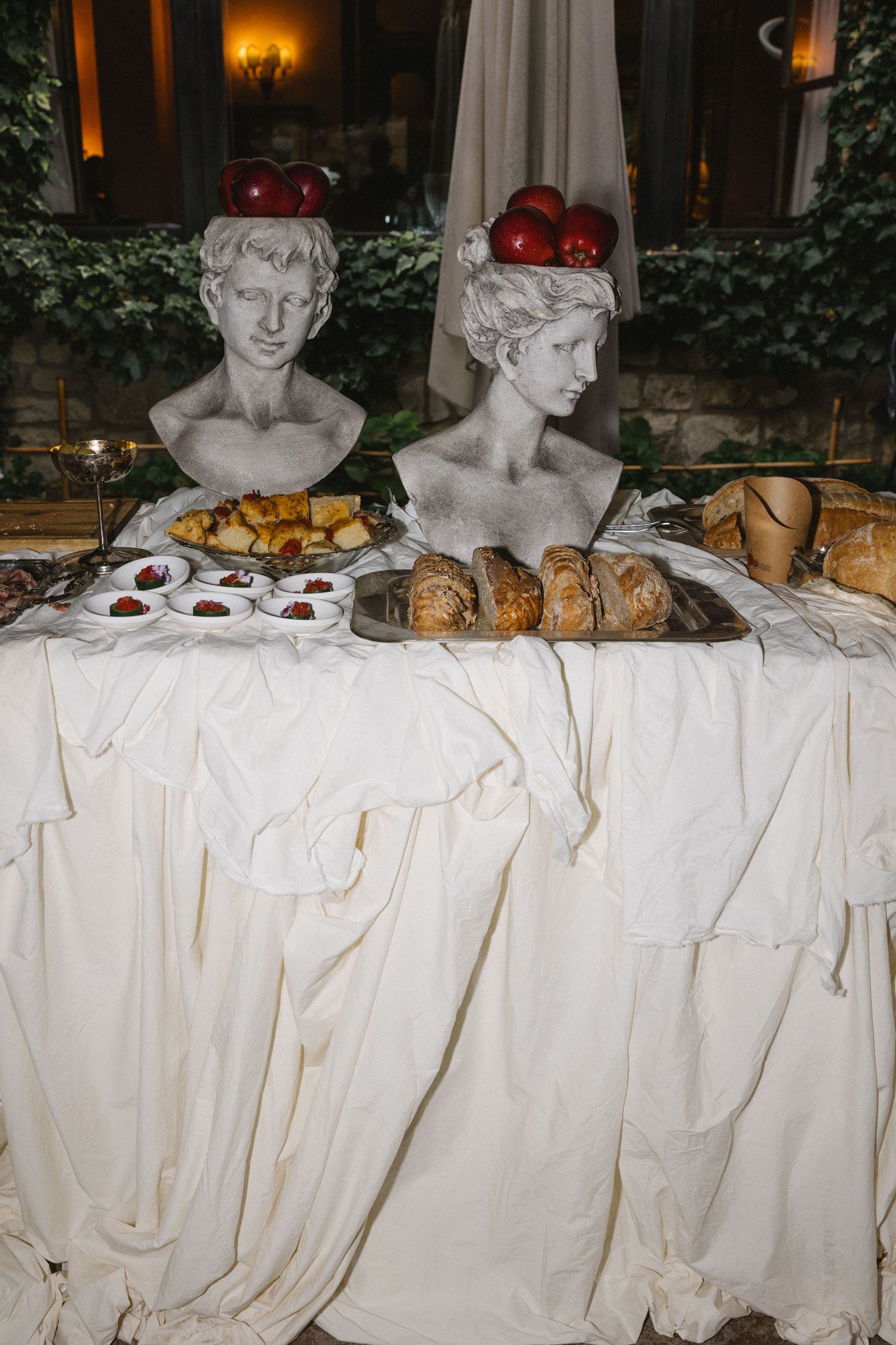Two classical busts with red apples on their heads displayed on a draped white table with bread and pastries.