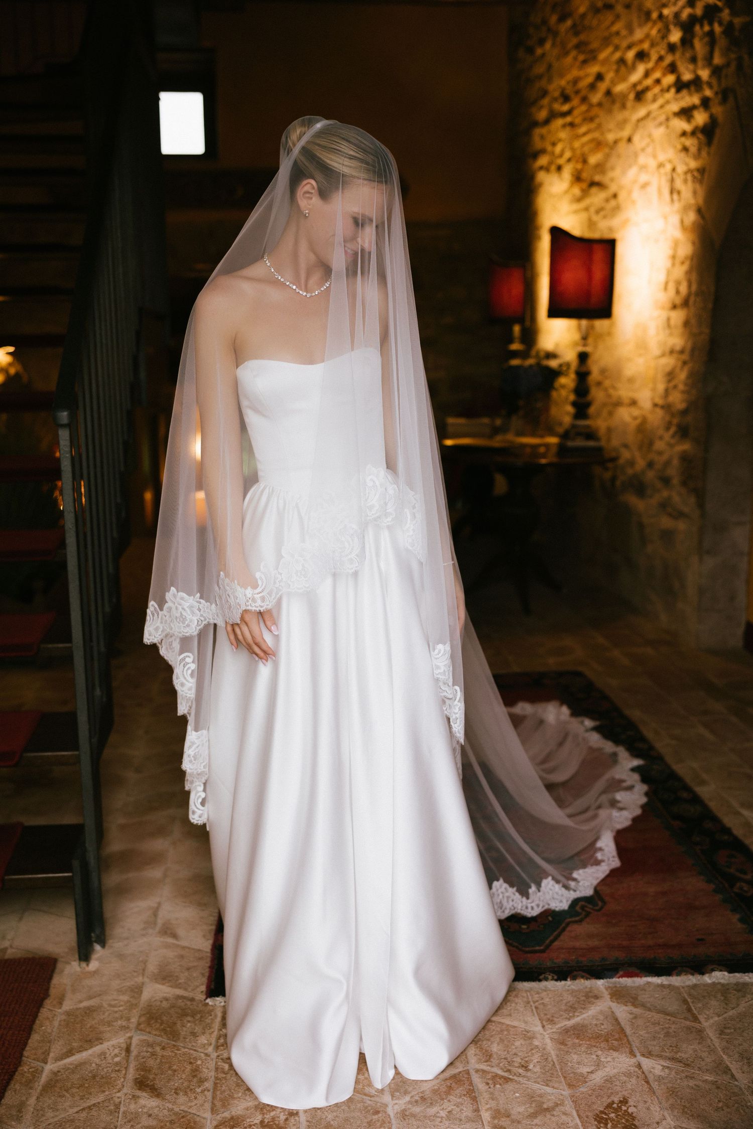 Bride in strapless white satin gown with lace-trimmed veil stands on tiled floor near staircase in warmly lit rustic venue.