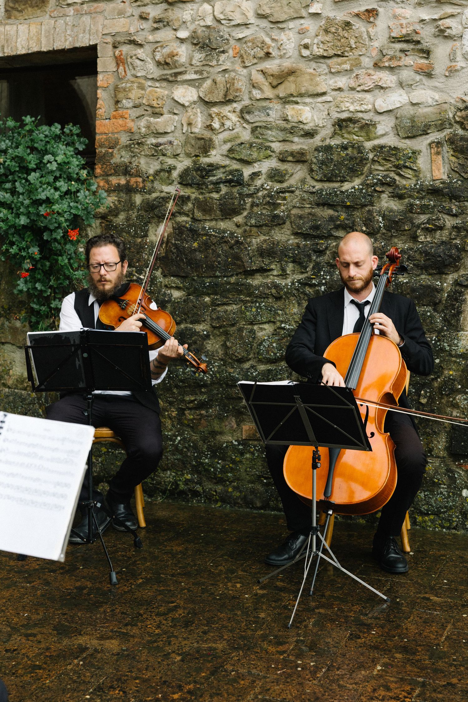 Two musicians playing violin and cello outdoors against a stone wall, reading sheet music on stands.