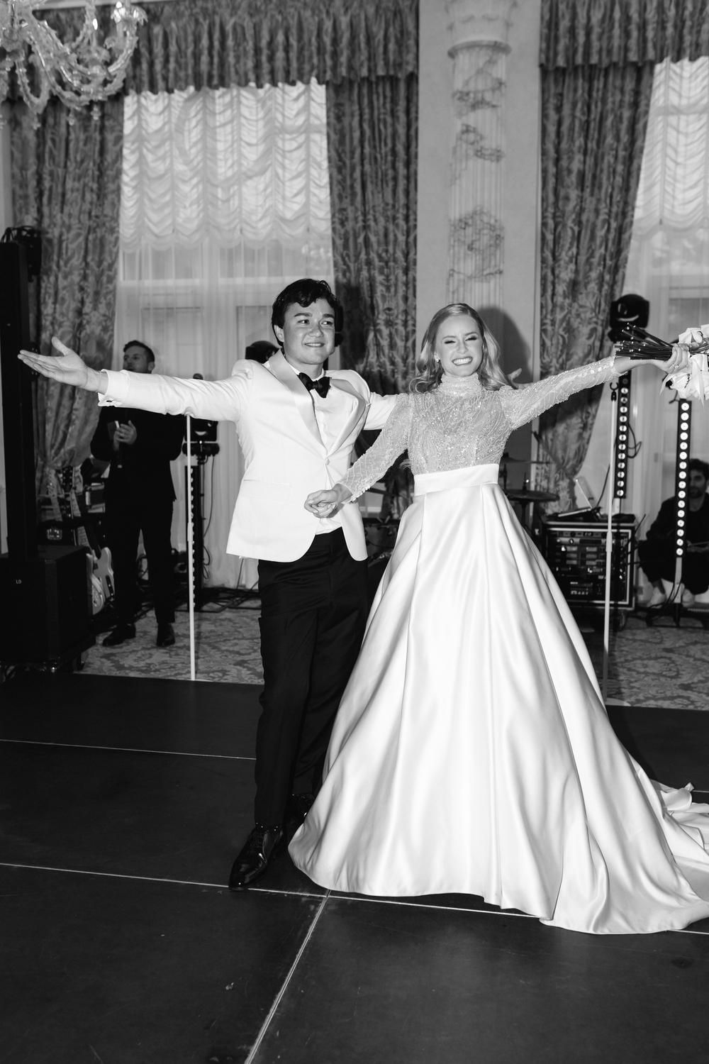 Black and white photo of a bride and groom dancing joyfully at their wedding reception in an elegant ballroom.