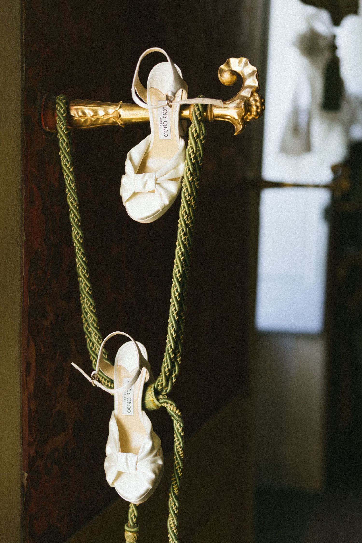White Jimmy Choo bridal heels hanging on a golden ornate hook with a green rope in a dimly lit room.