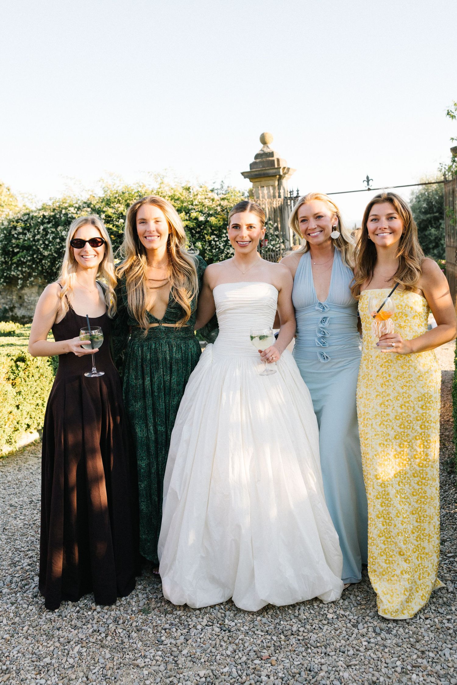 Bride in white ball gown posing with four female guests in colorful formal dresses at an outdoor wedding.