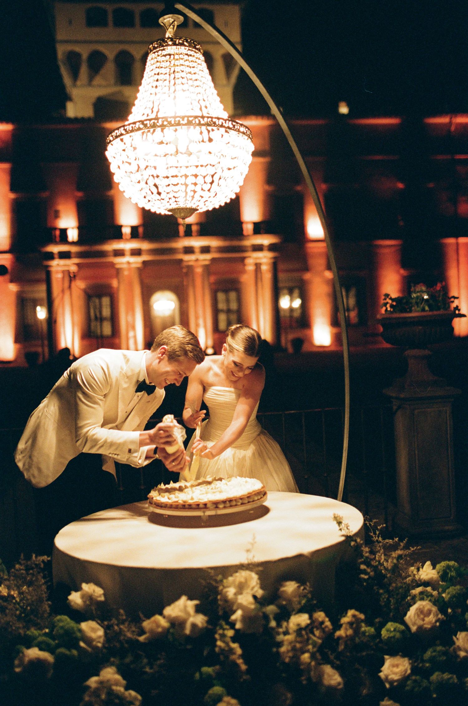 Bride and groom cutting wedding cake under crystal chandelier with romantic amber lighting at outdoor evening reception.