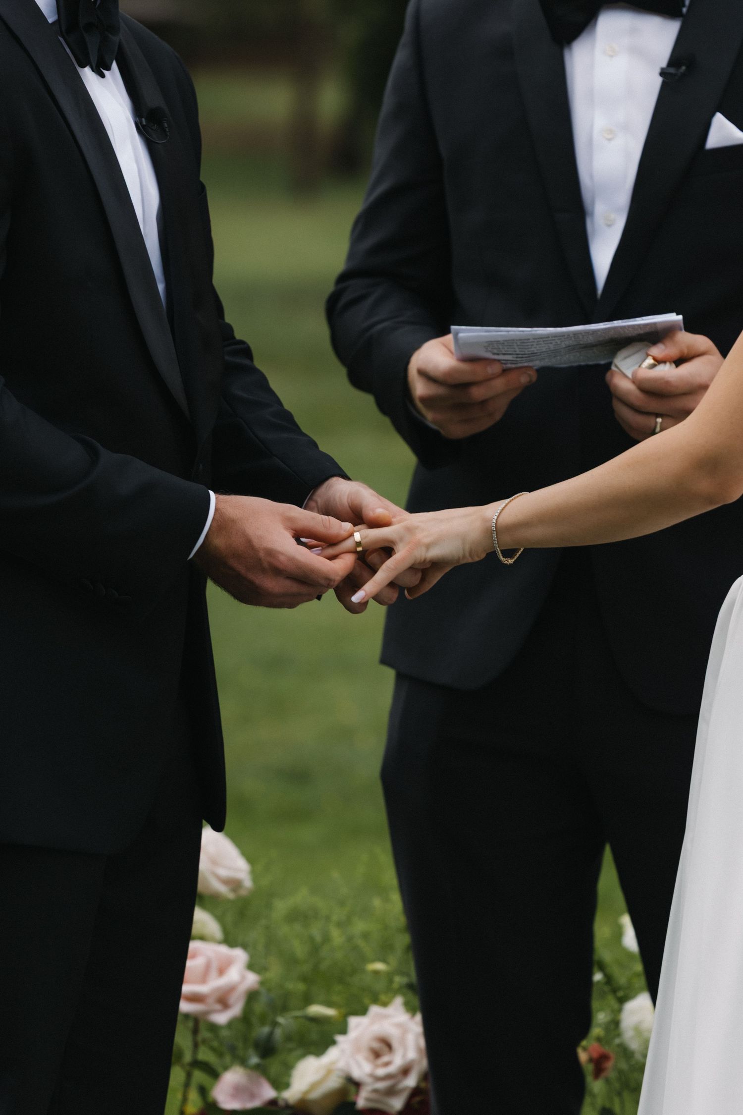 Groom placing a gold wedding ring on bride's finger during an outdoor ceremony, with officiant holding vows in background.