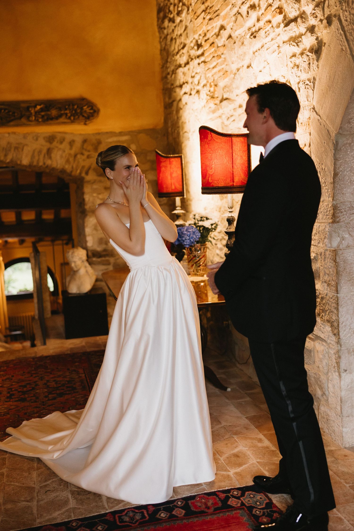 Bride covers her mouth in emotion during first look with groom in rustic stone venue with warm lighting.