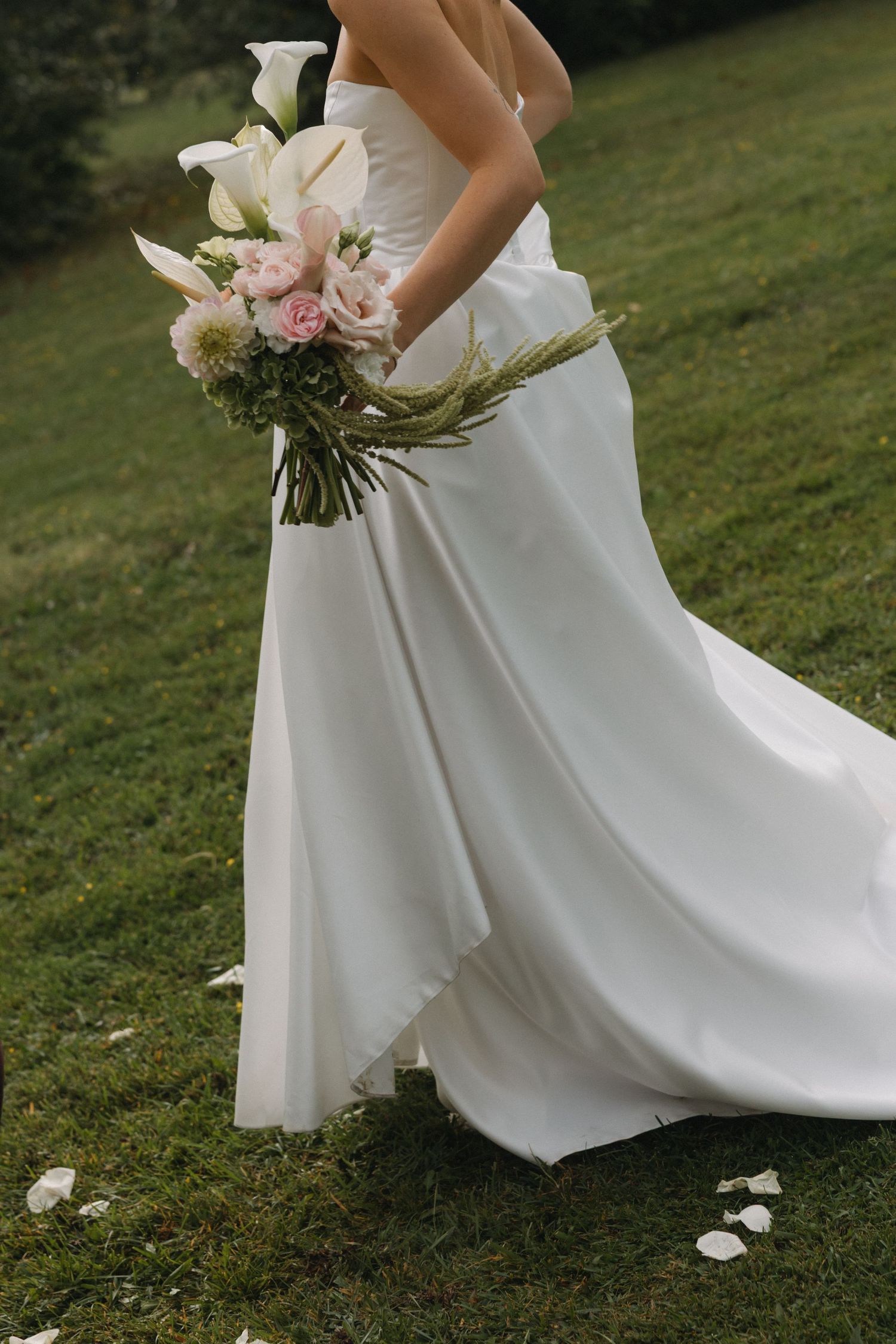 Bride in white gown holding bouquet of calla lilies and pink roses on green grass.
