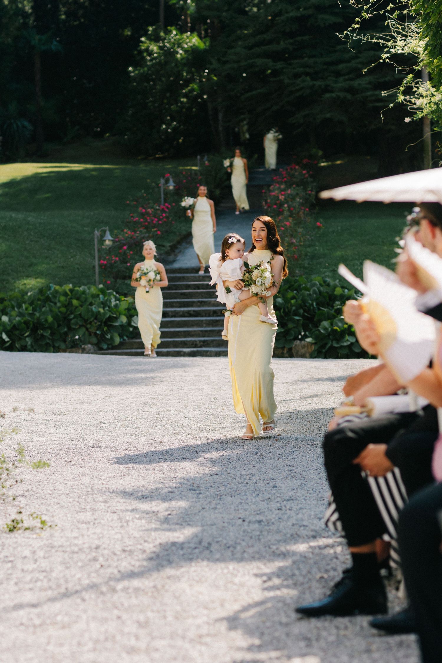 Bridesmaids in yellow dresses walk down garden path carrying bouquets at outdoor wedding ceremony.