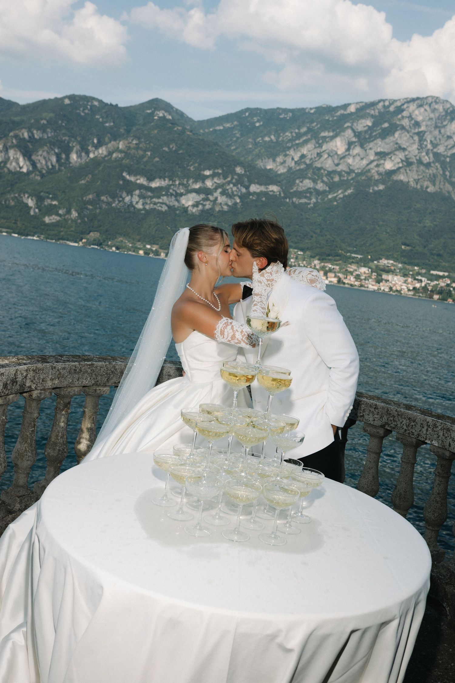 Bride and groom kissing behind champagne tower on balcony overlooking scenic lake and mountains on wedding day.