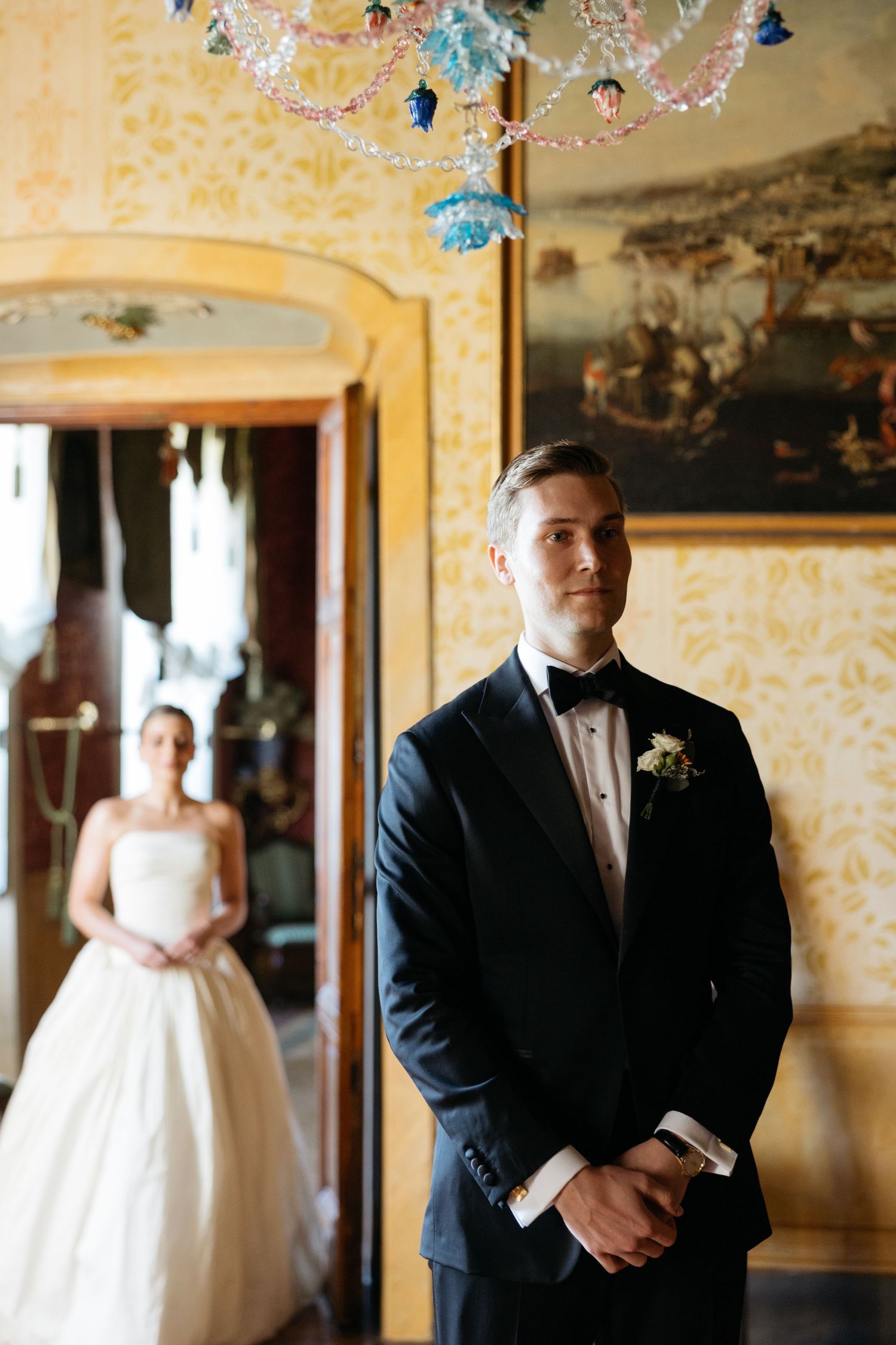 Groom in black tuxedo with boutonniere stands waiting as bride appears in doorway during wedding first look.